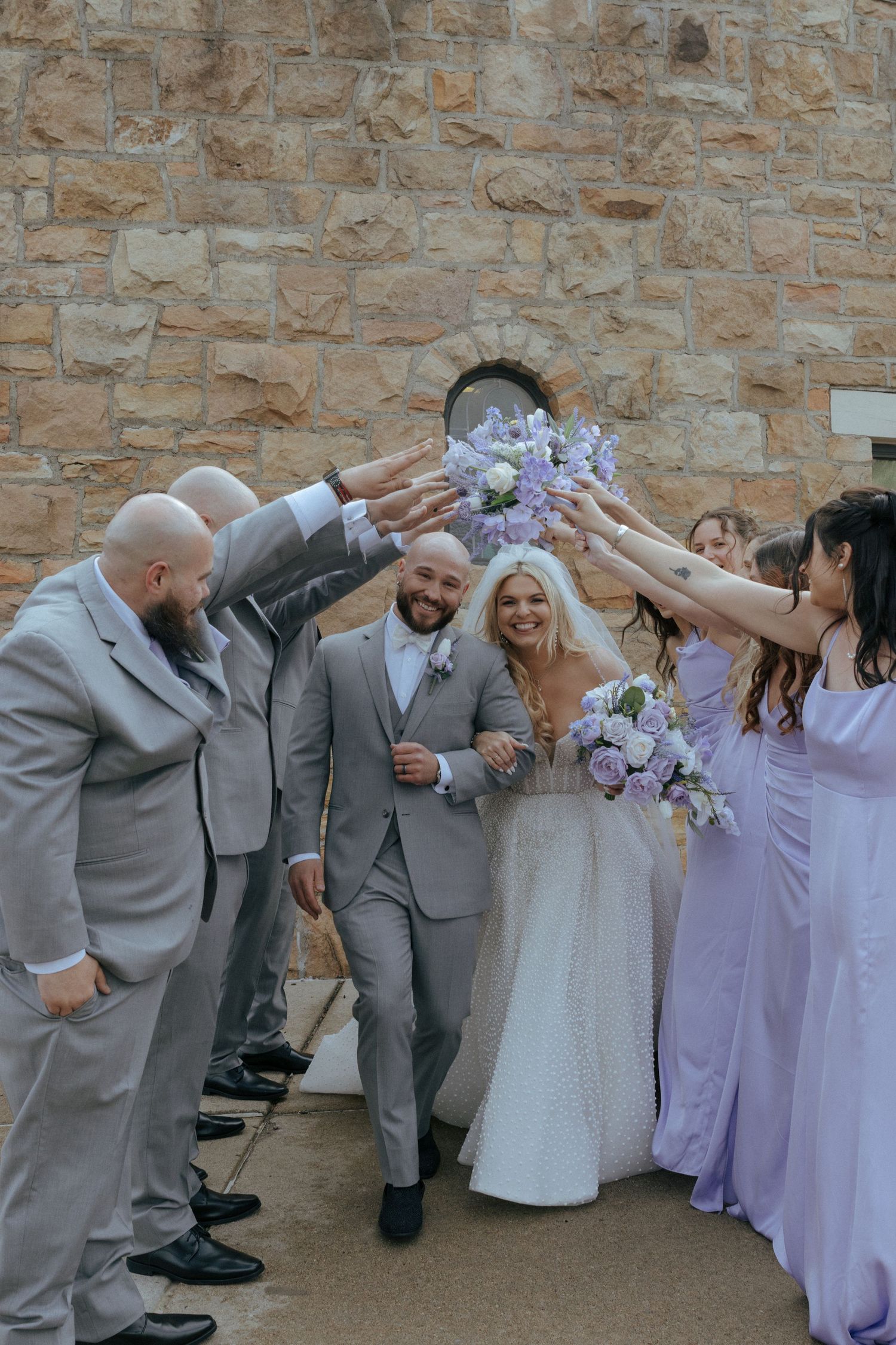 Wedding party in gray suits and lavender dresses performs synchronized dabbing dance move against stone wall.
