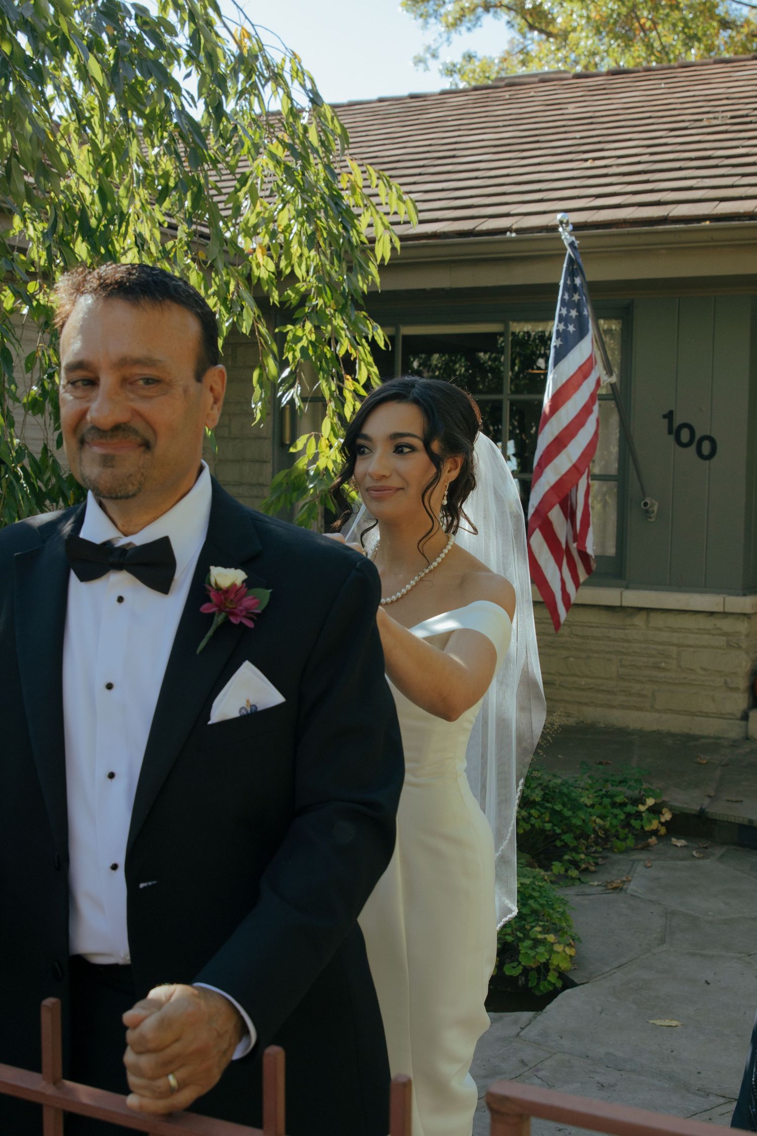 A couple in formal wedding attire share a joyful moment together outdoors with an American flag in the background.