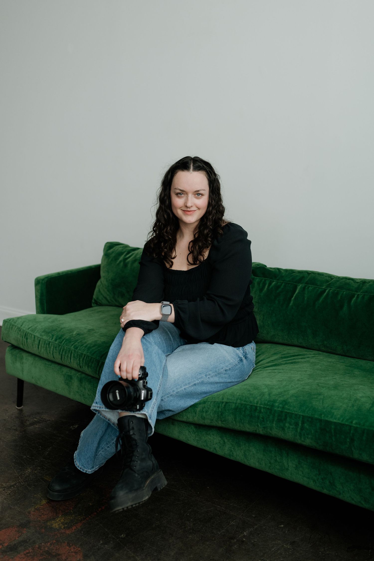A person in black attire reclines on an emerald green velvet sofa against a light gray wall.