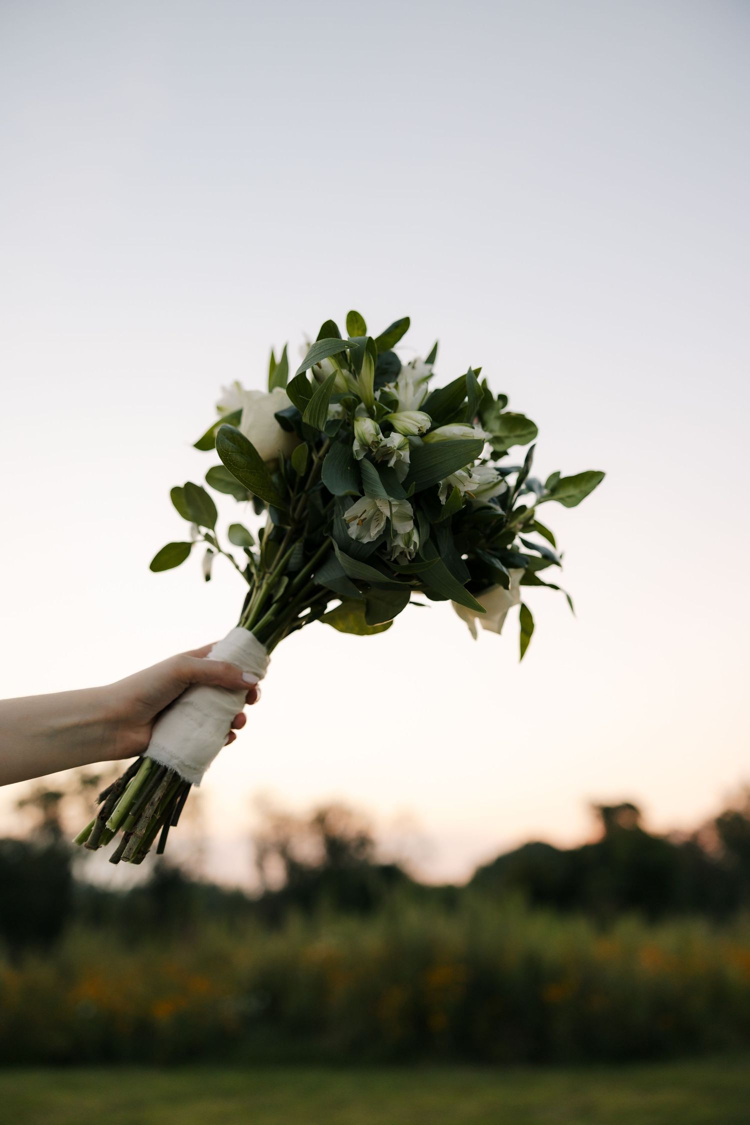 Hand holding a white and green bridal bouquet with eucalyptus and white flowers against a twilight sky.