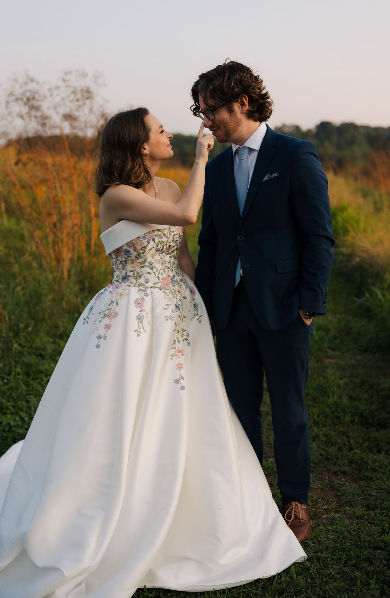 Bride in floral embroidered ball gown touches grooms face outdoors at golden hour.