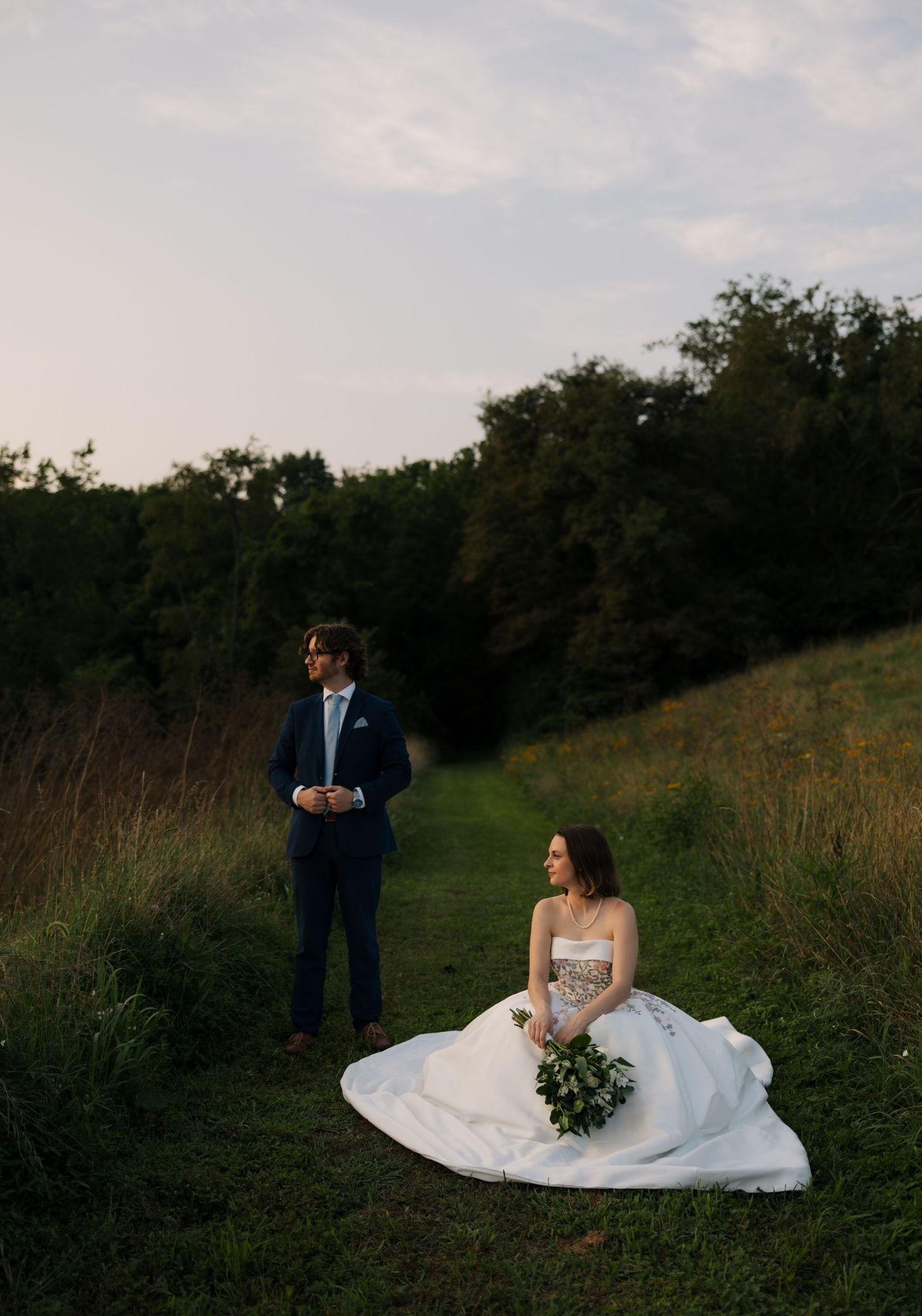 Bride in floral wedding gown sits on grass while groom stands on scenic green hillside at sunset.