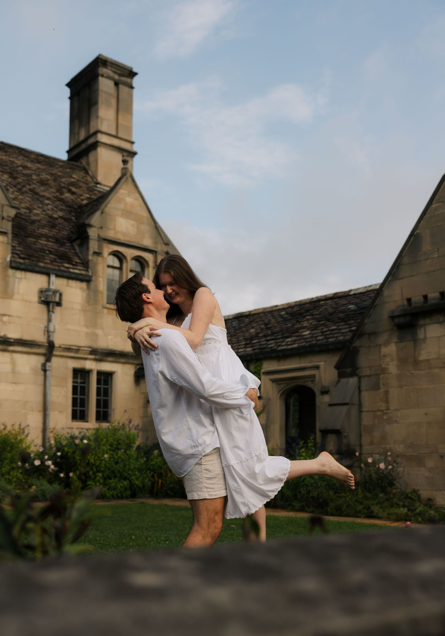 A couple shares a romantic moment as a man lifts a woman in white outside a historic stone manor.
