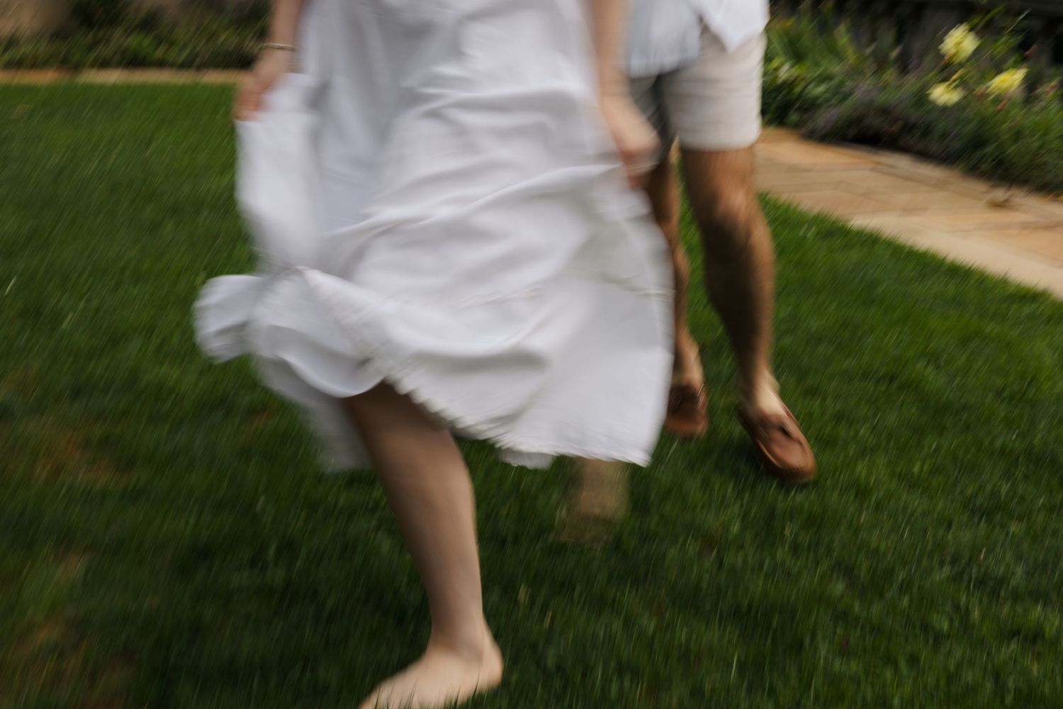 Blurred motion shot of couple running barefoot through green grass in white outfits.
