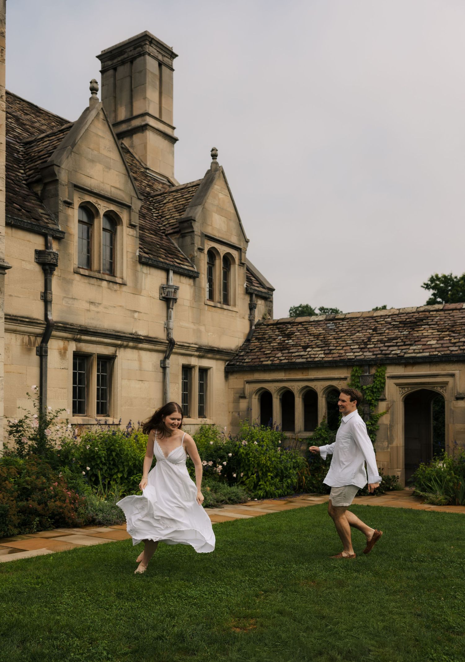Joyful couple in white outfits running and playing on the lawn of a grand historic stone manor house.