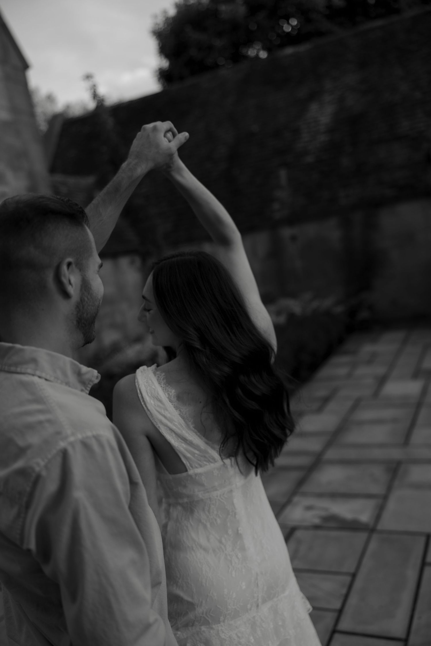 Black and white photo of a couple dancing outdoors, man spinning woman with raised hands near a stone wall.
