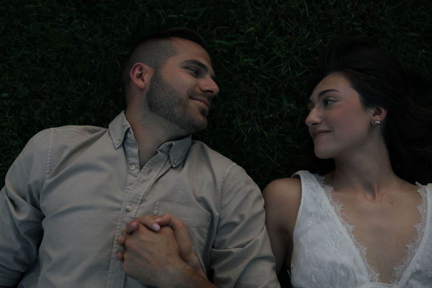Couple lies on green grass holding hands and gazing at each other during engagement session.