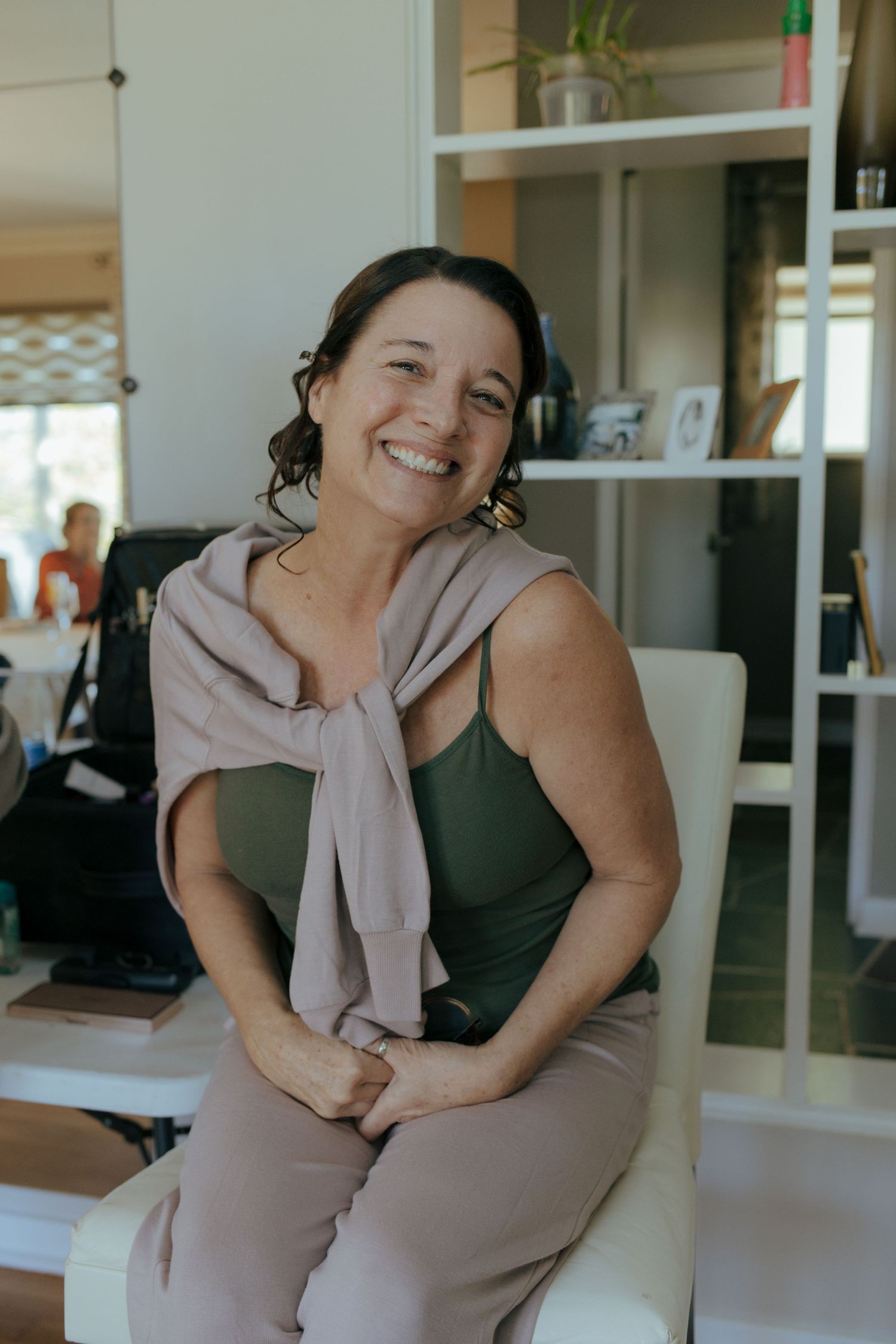 Smiling woman in olive green dress with taupe wrap sits in white chair in bright modern office space.