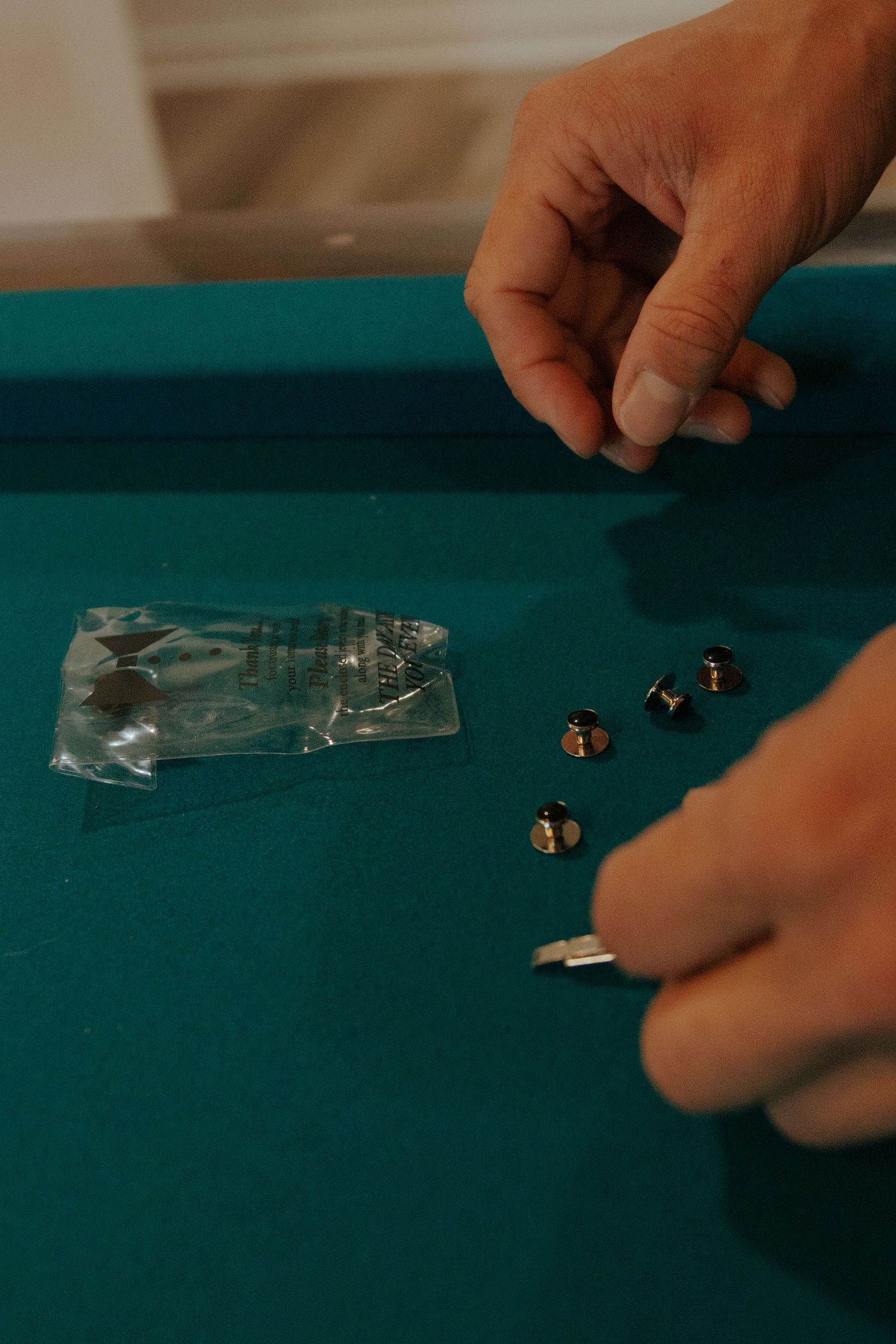 Hands arranging small silver tuxedo studs on a green felt surface next to an open plastic packaging bag.