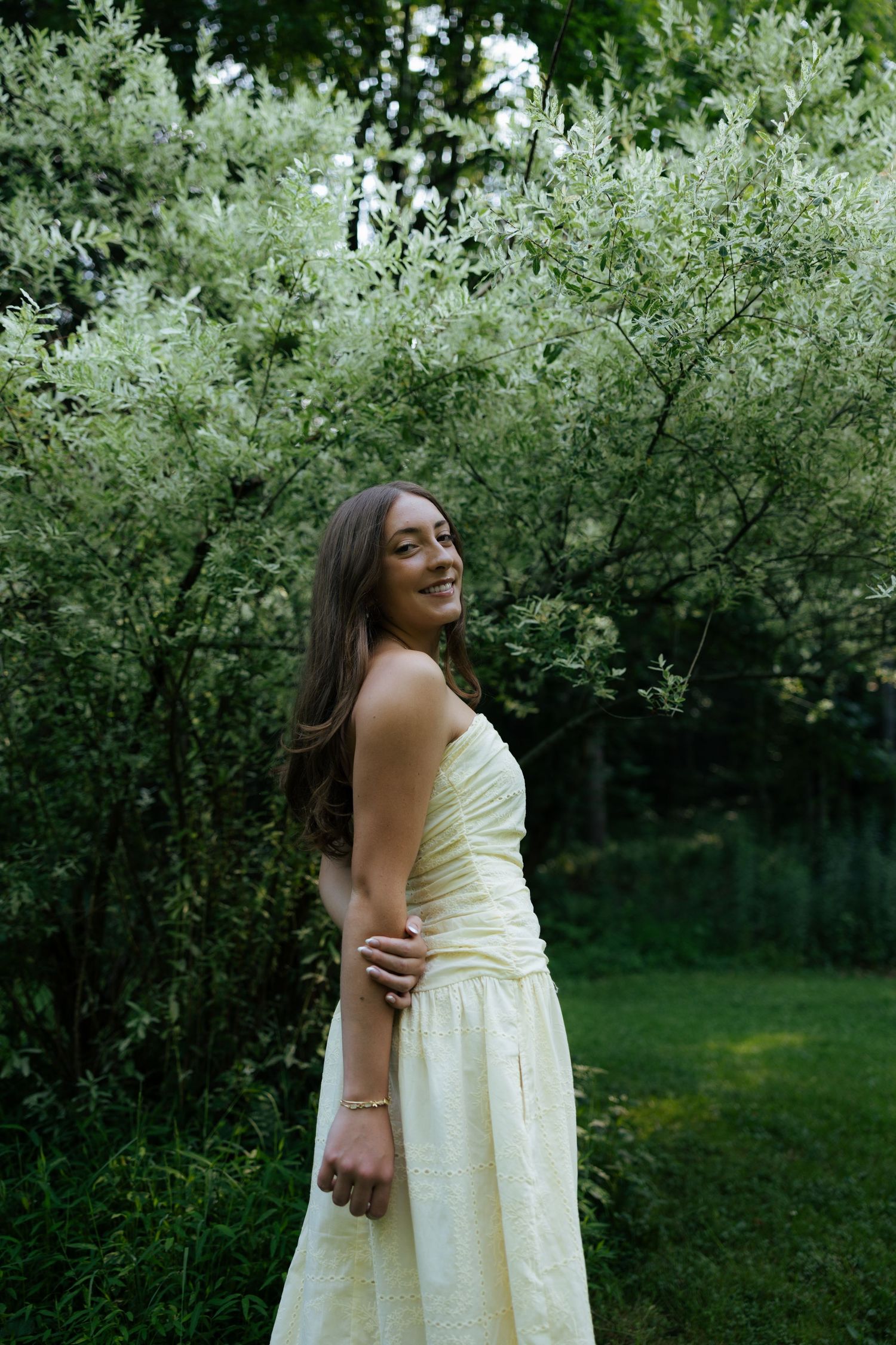 Young woman in a pale yellow strapless dress smiling beside blooming white flowering shrubs outdoors.