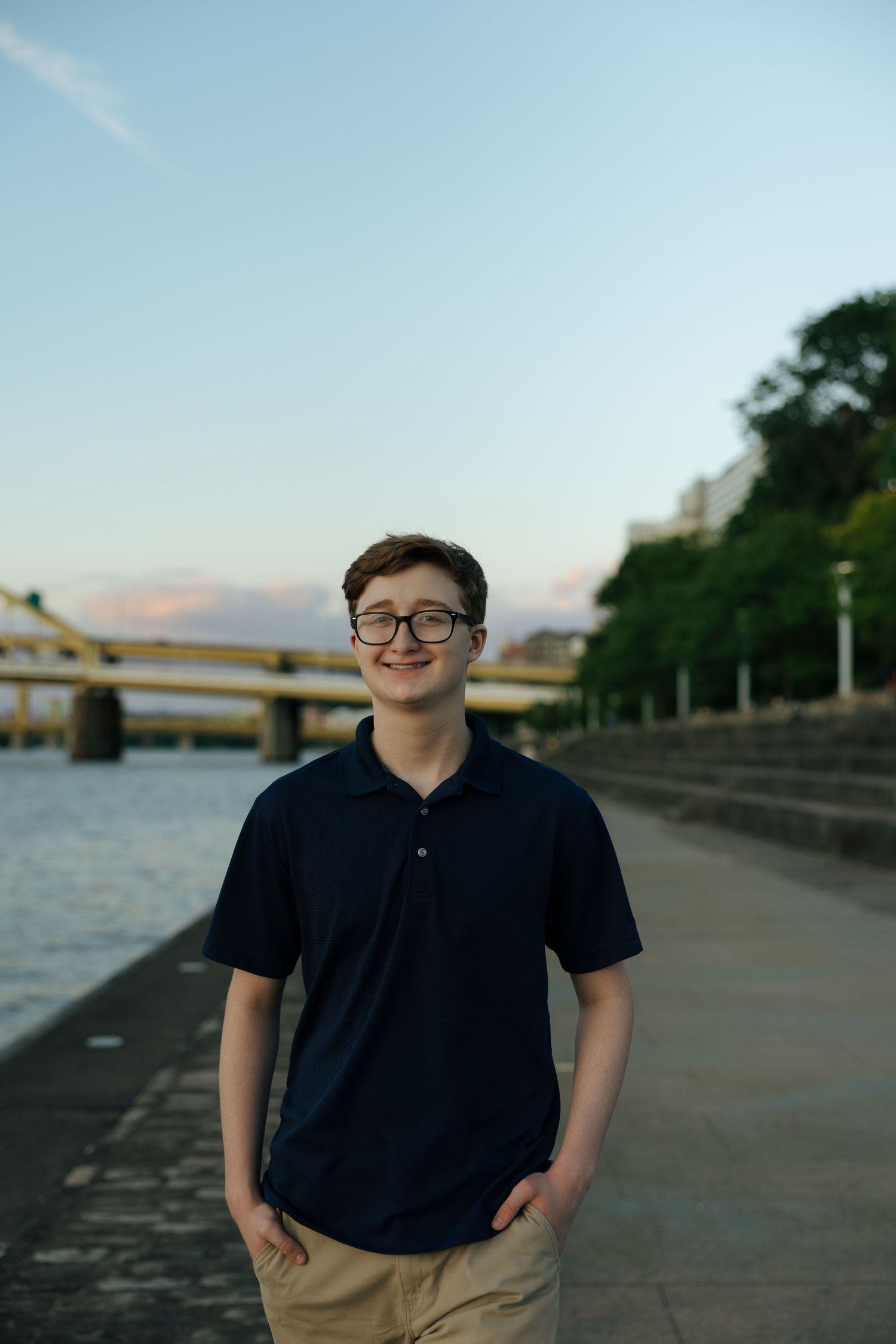 Smiling teen boy in navy polo and khakis standing along Pittsburgh riverfront walkway at dusk.