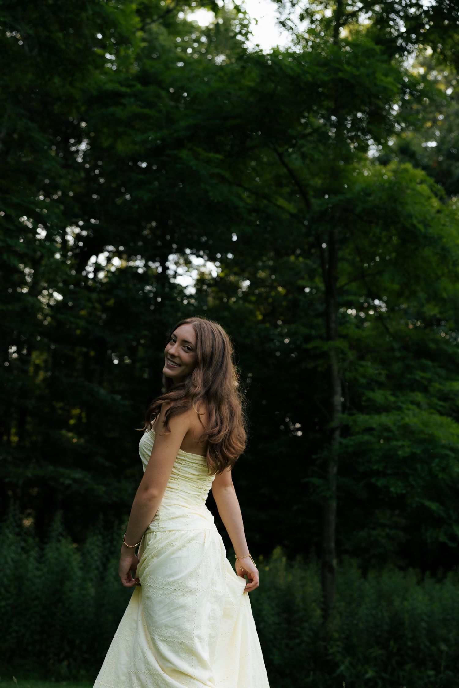 Smiling bride in white strapless gown twirling in lush green forest setting.