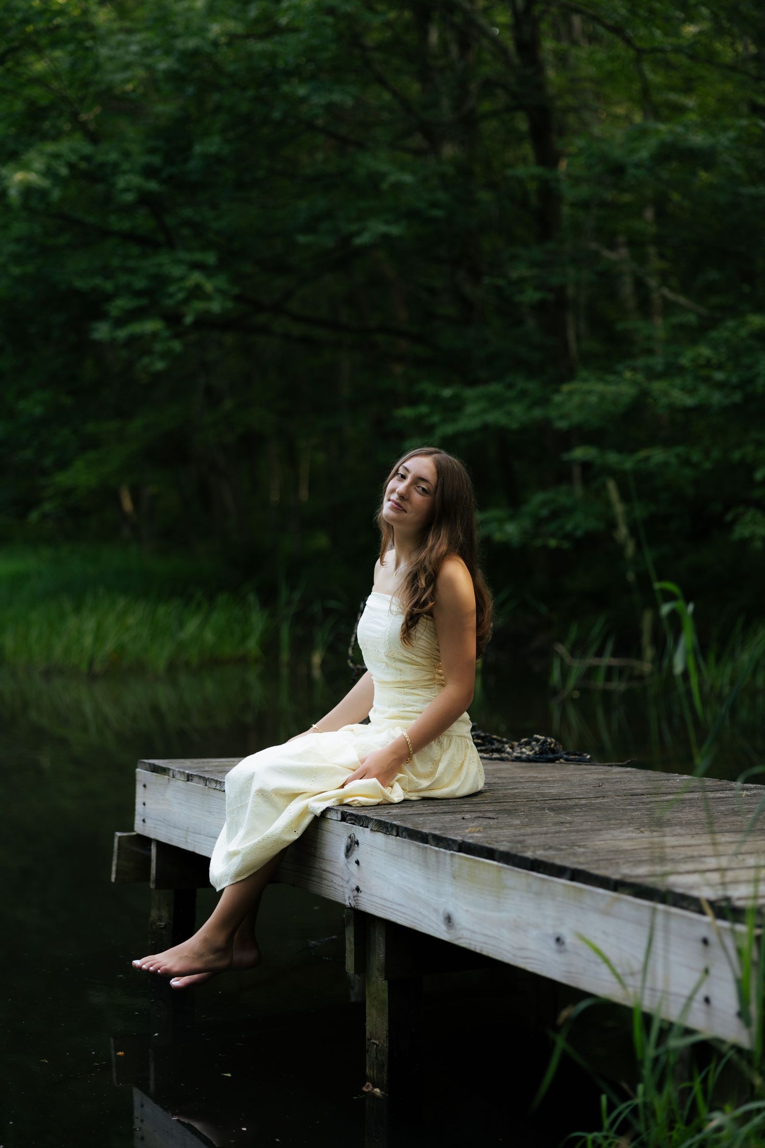 Young woman in yellow dress sitting barefoot on a wooden dock by a calm forest lake at dusk.