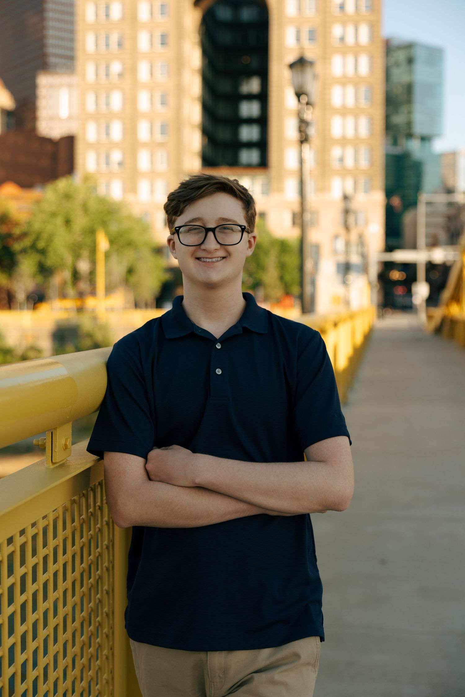 Smiling teen boy with glasses and crossed arms standing on yellow bridge with city buildings behind.