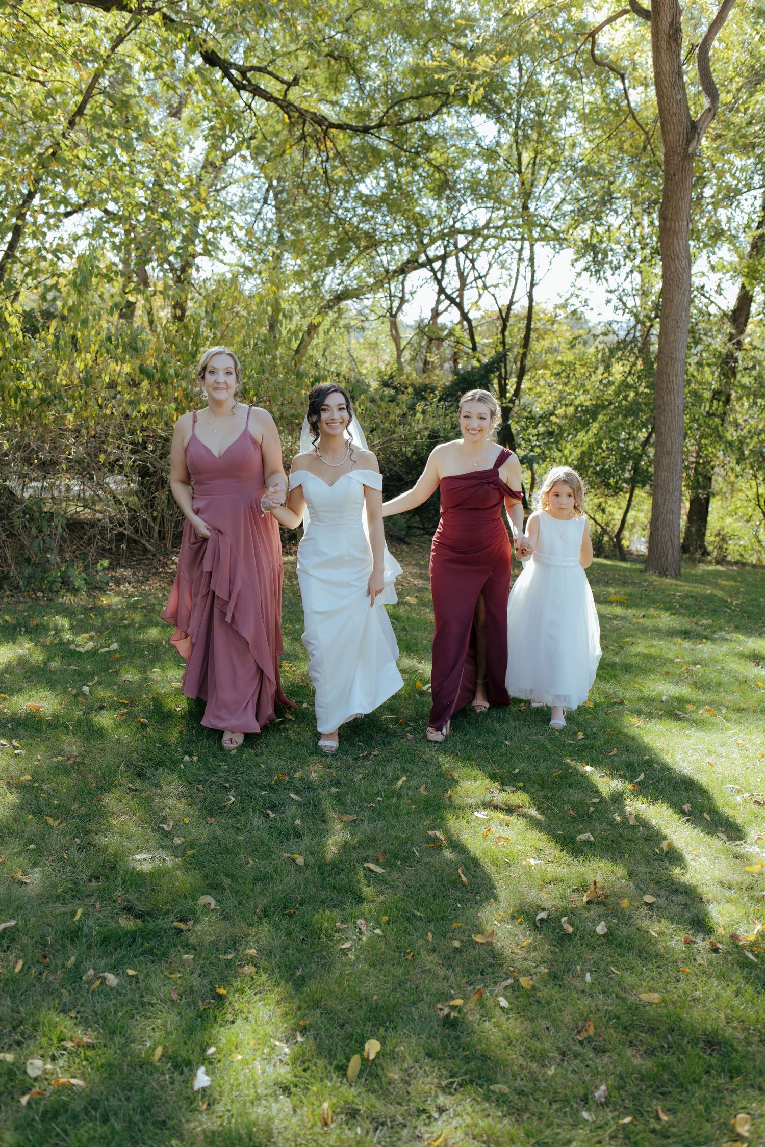 Bride in white gown walks with bridesmaids in burgundy and mauve dresses and flower girl through a green garden.