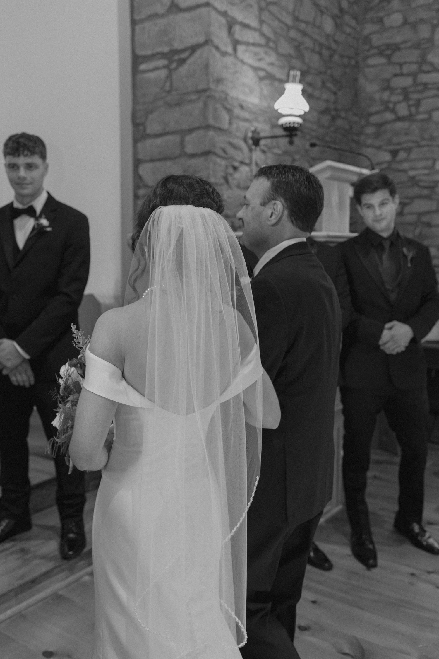 Black and white photo of bride and groom exchanging vows at altar with groomsmen standing nearby in stone church.
