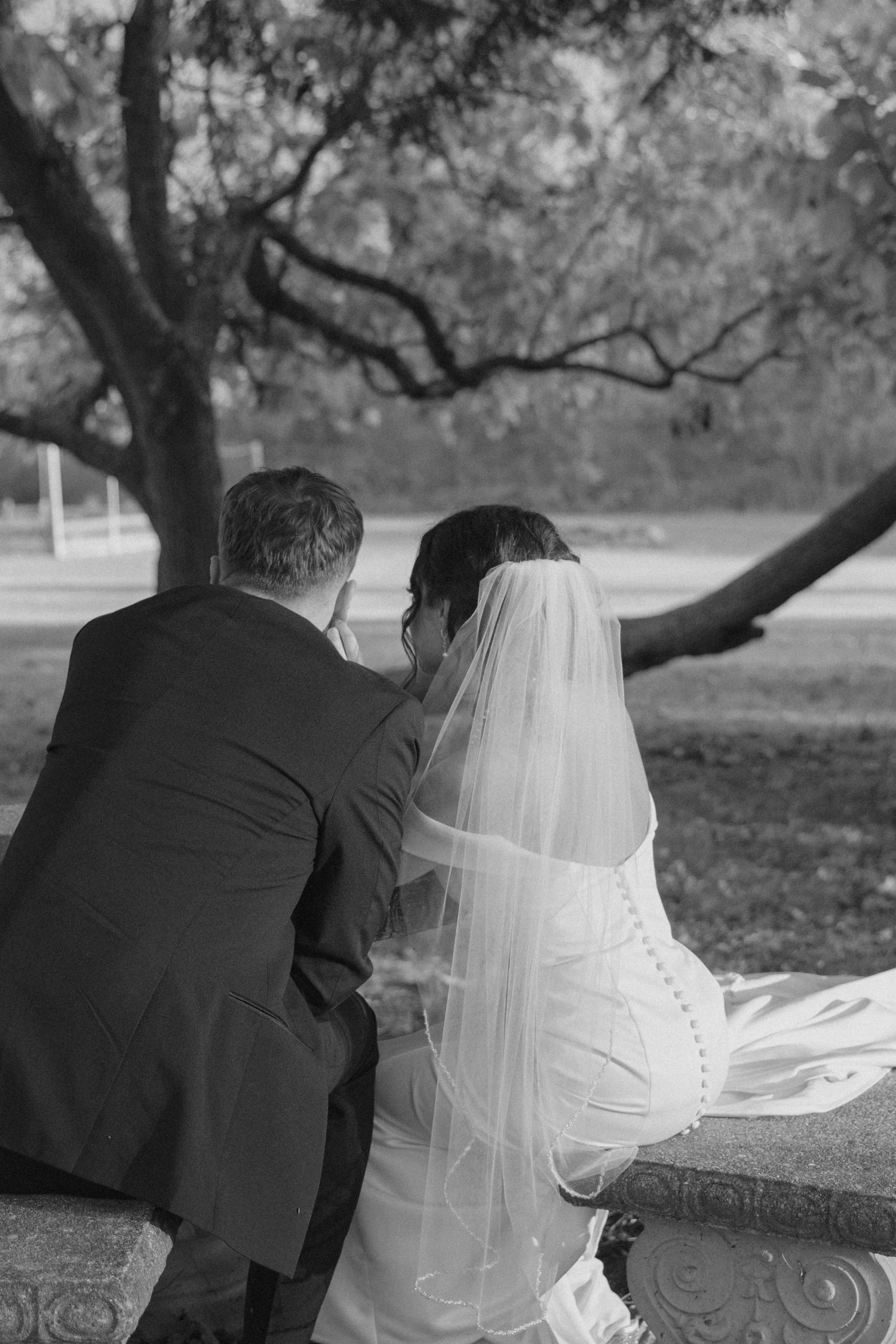Black and white photo of bride and groom sitting together on a stone bench beneath a large tree.