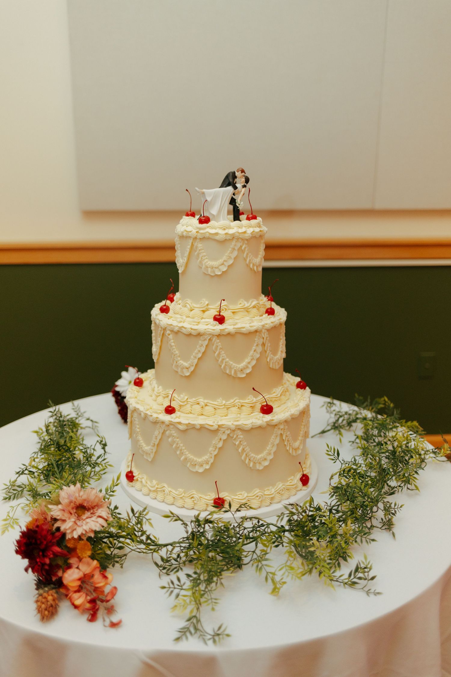 Elegant four-tier white wedding cake with cherry decorations, piped frosting swags, and bride and groom topper on a white table.