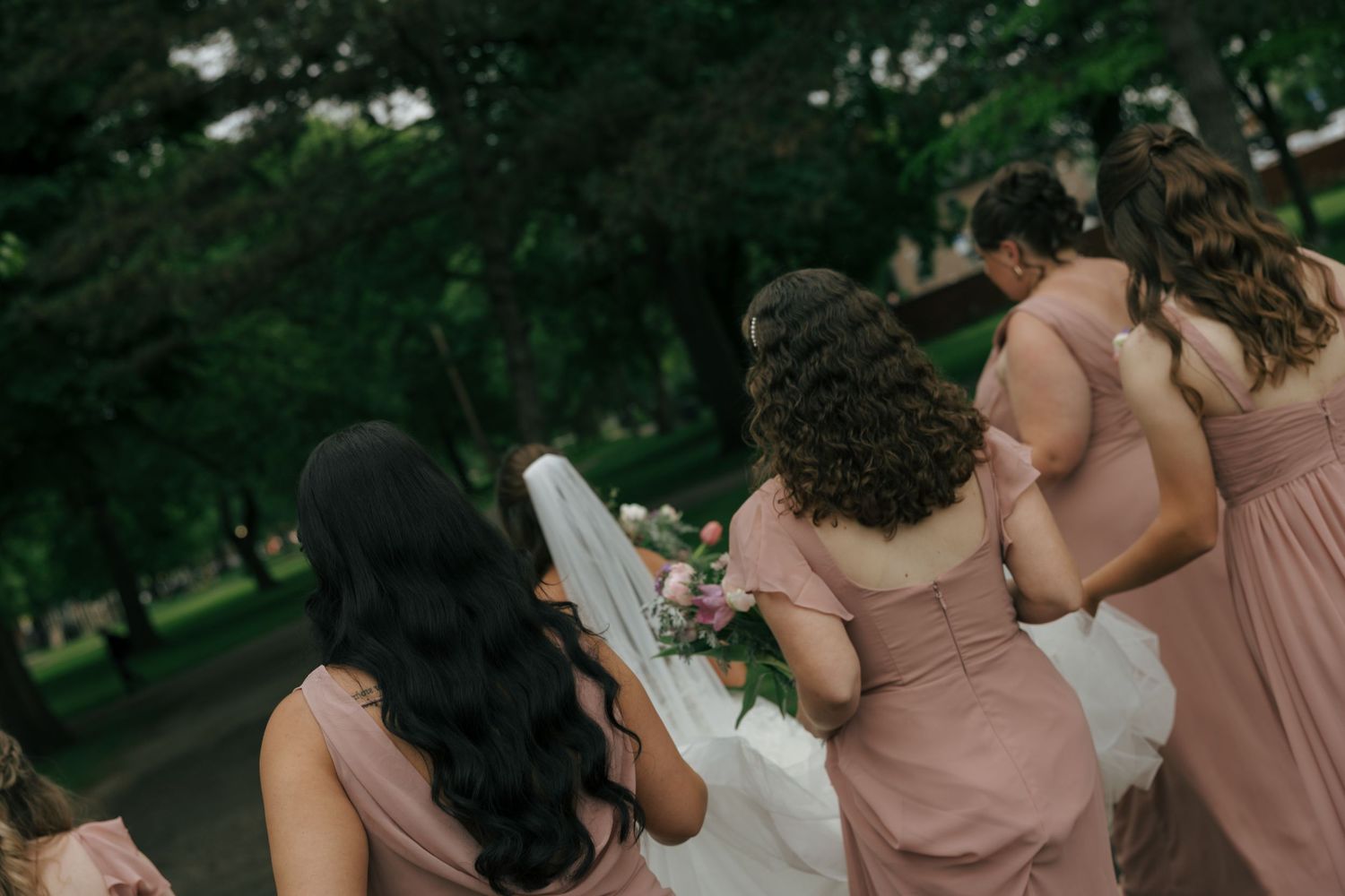 Bridesmaids in dusty rose dresses surround a bride outdoors, adjusting her white veil and bouquet.