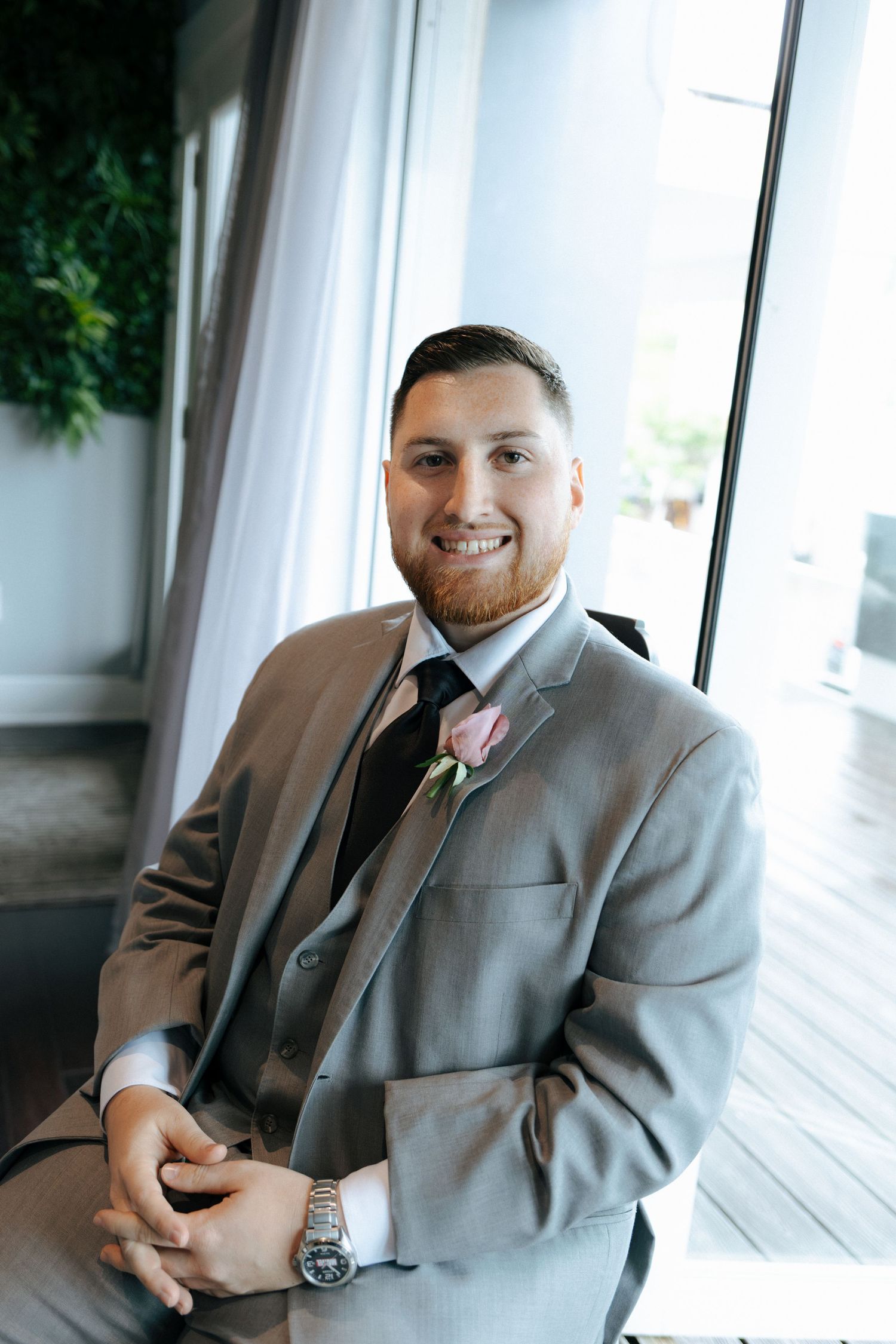 Smiling groom in gray suit with pink boutonniere and black tie seated near window at wedding venue.