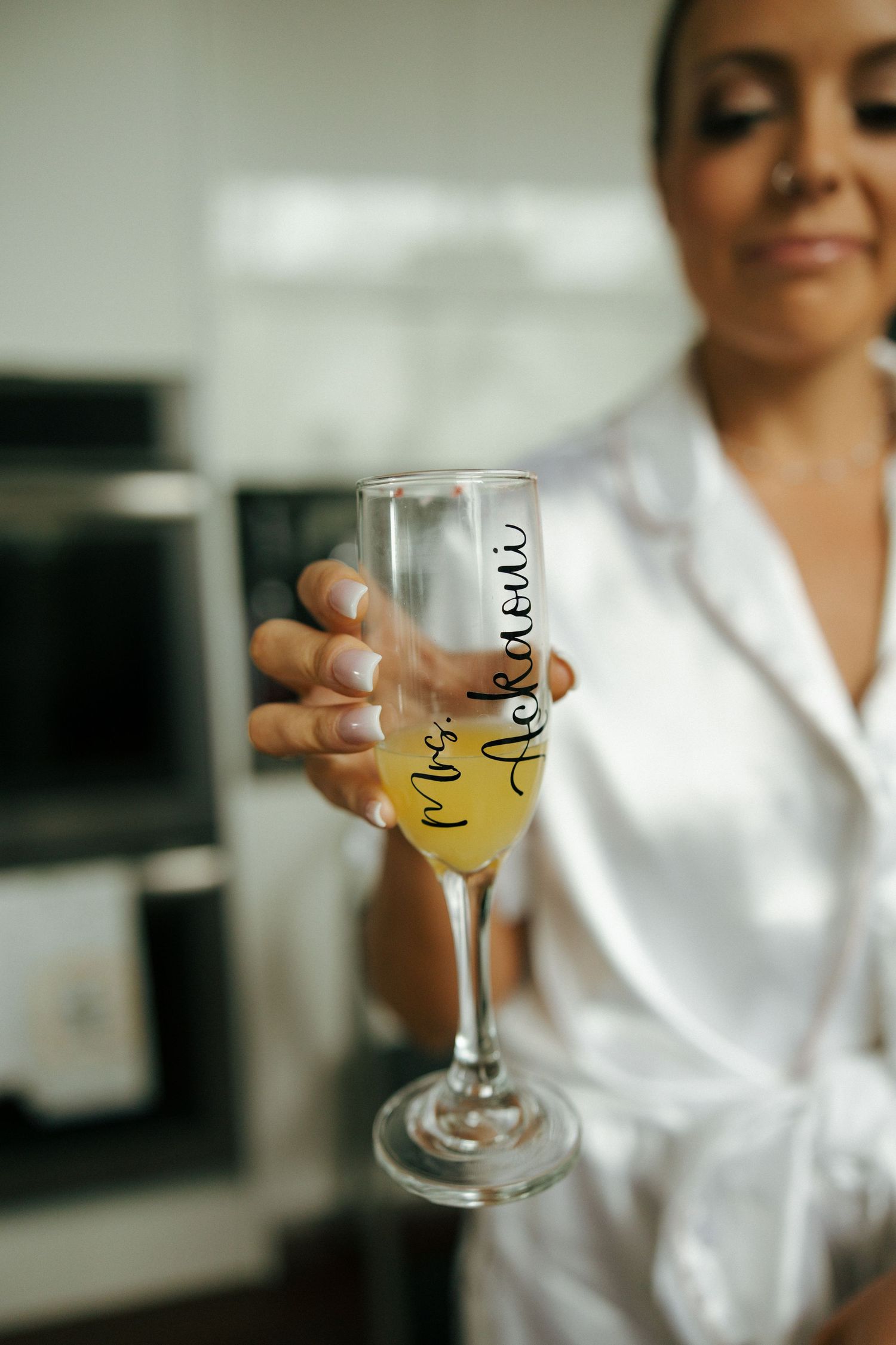 Bride in white robe holding personalized Mrs. champagne flute with yellow bubbly on wedding morning.