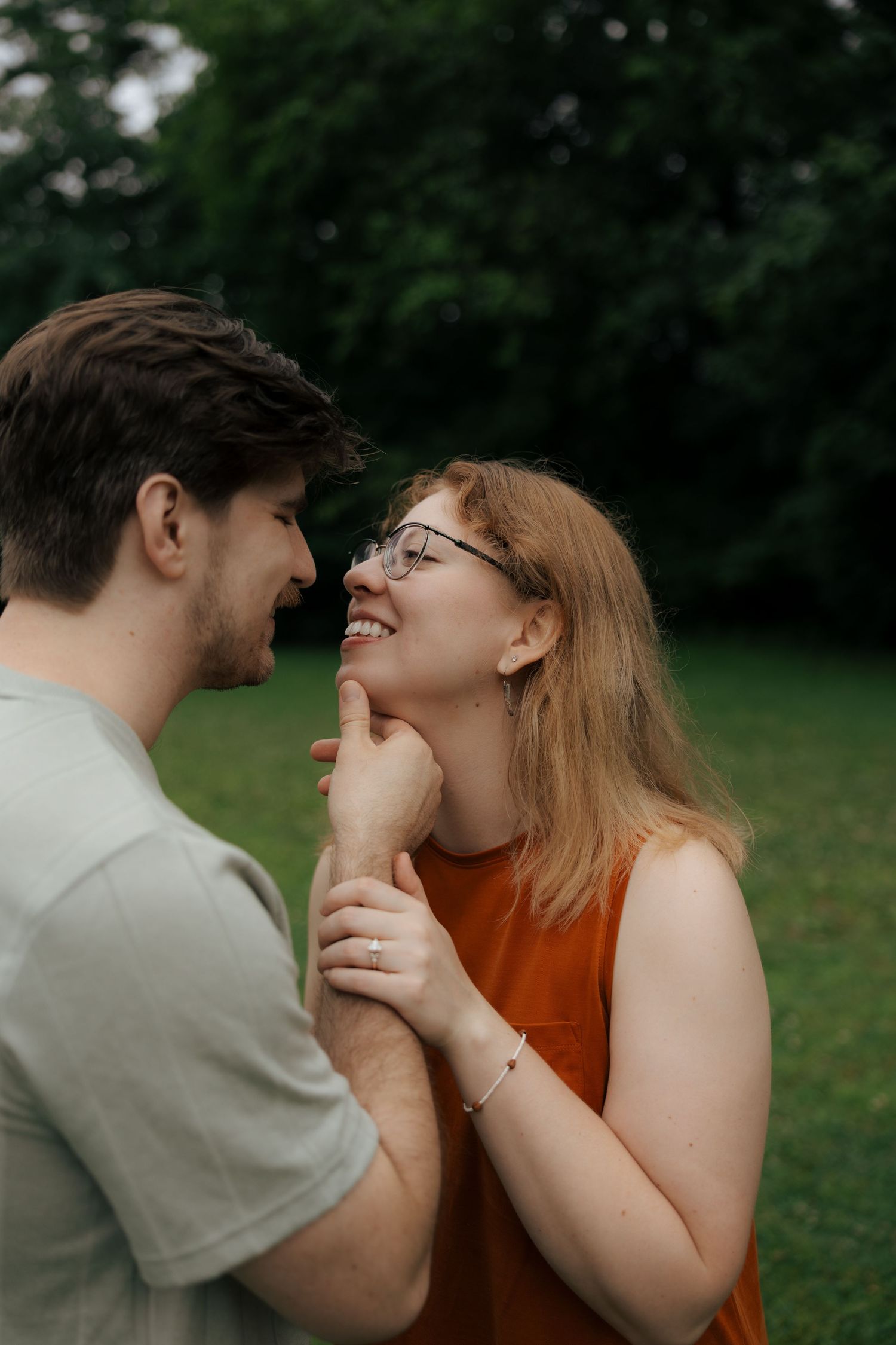 Smiling engaged couple holding hands close together in a green park setting.