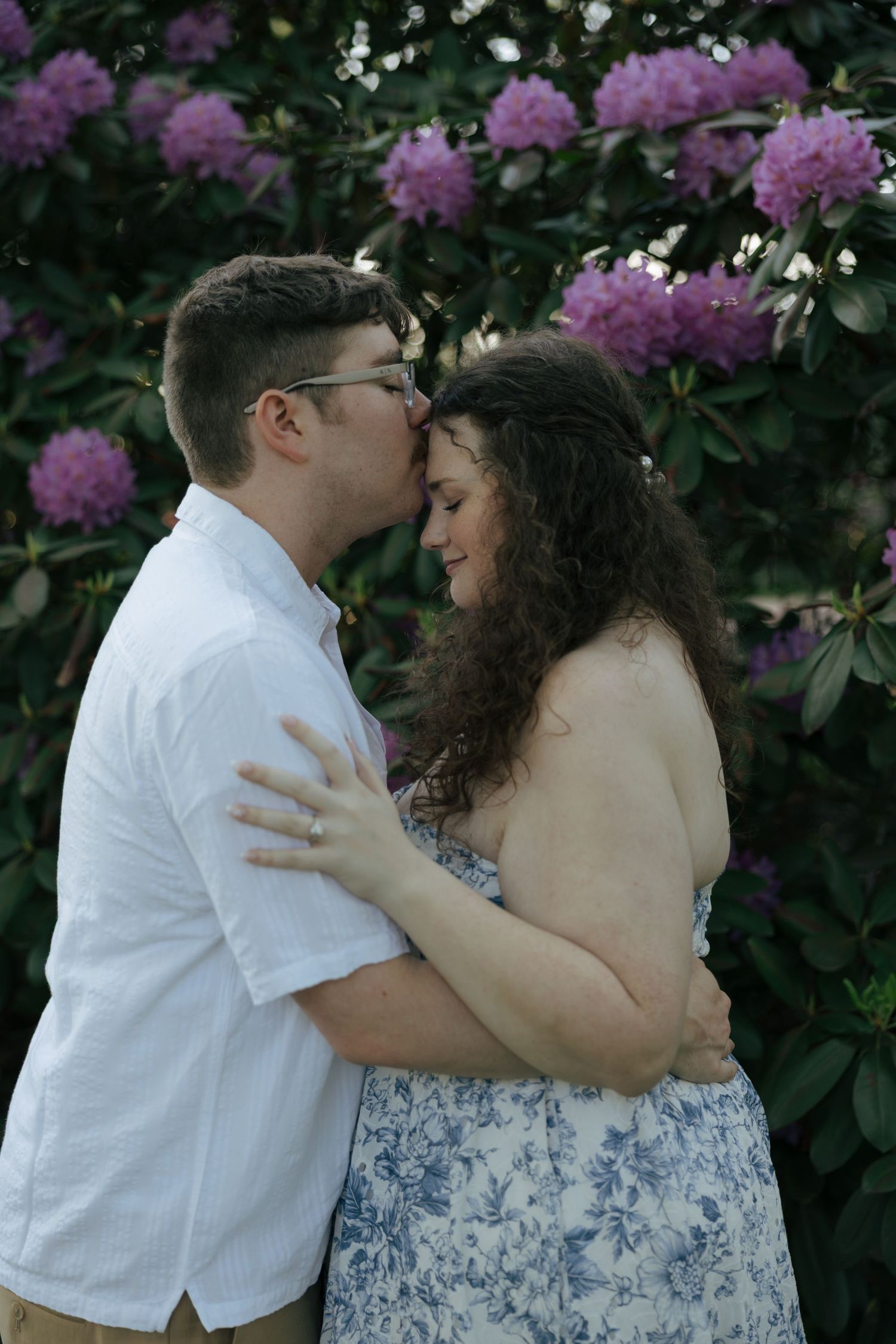 Engaged couple embracing and kissing near blooming purple rhododendron bushes during golden hour outdoor shoot.