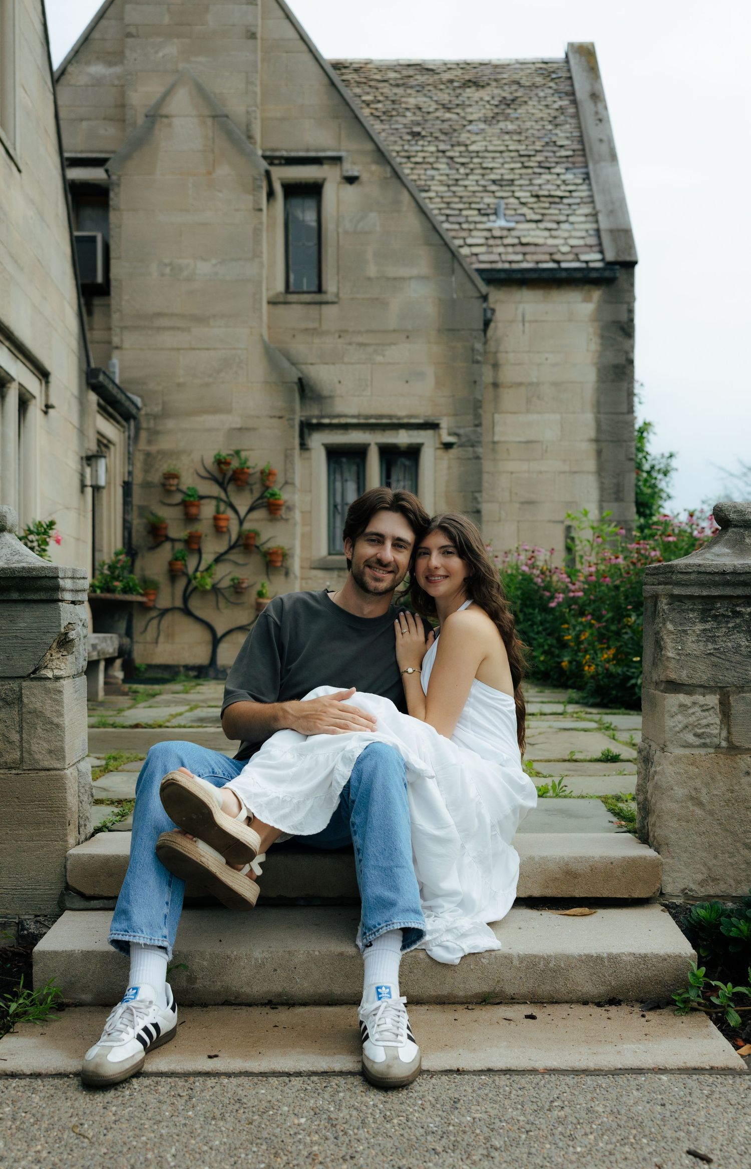 Smiling engaged couple sitting on stone steps in front of a charming stone cottage.