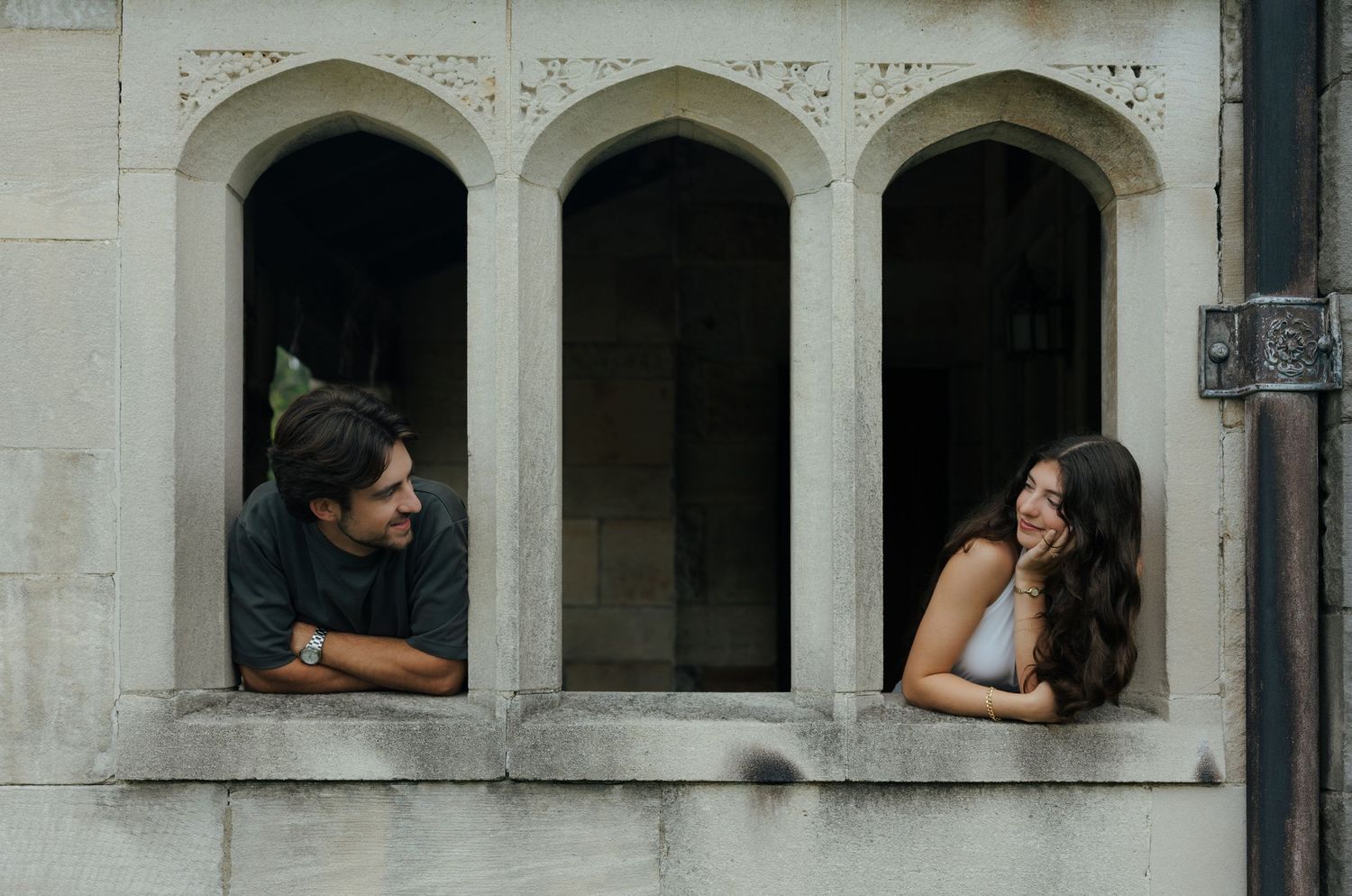 Couple leaning out of gothic stone arch windows smiling at each other during romantic engagement session.
