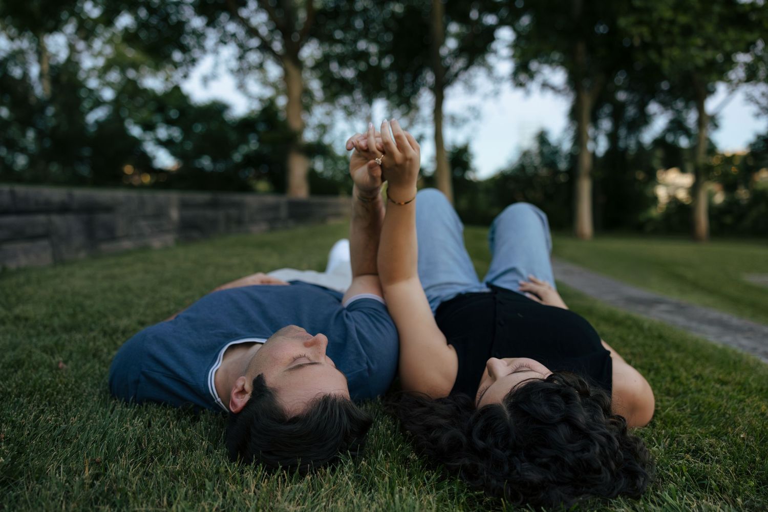 Couple lying on green grass in a park, holding hands and gazing at each other on a warm summer evening.