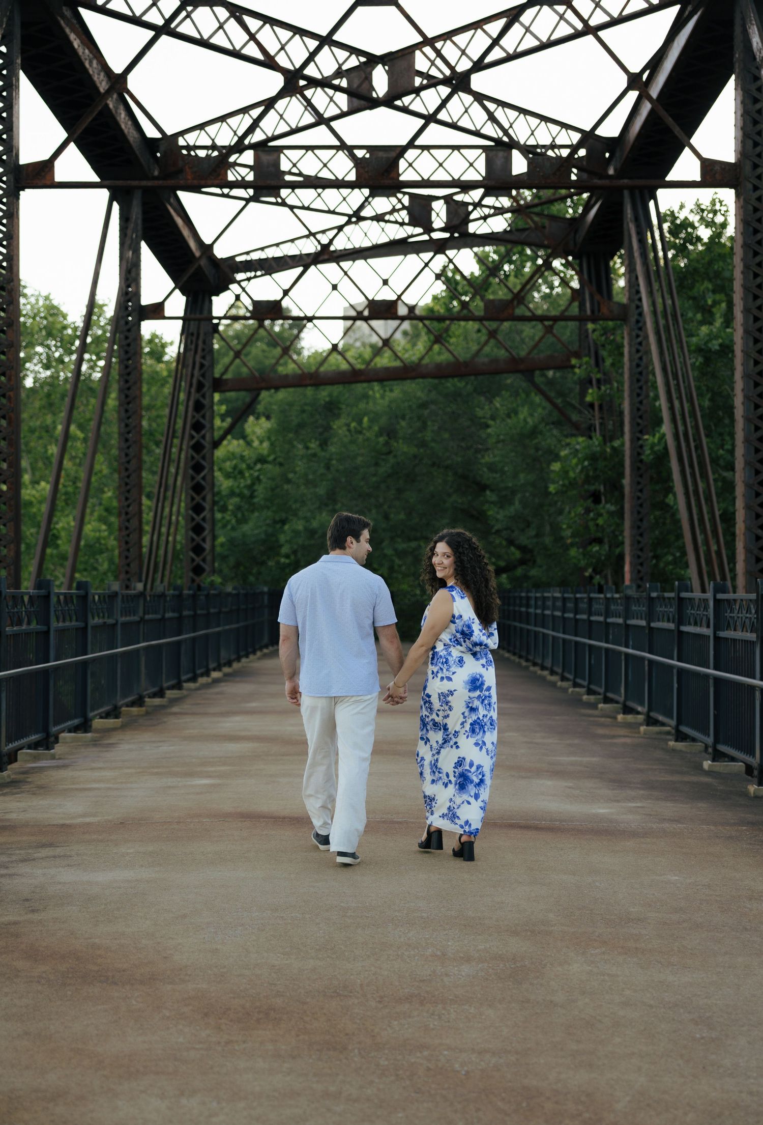 A couple walks hand in hand away from the camera along a scenic historic iron pedestrian bridge.