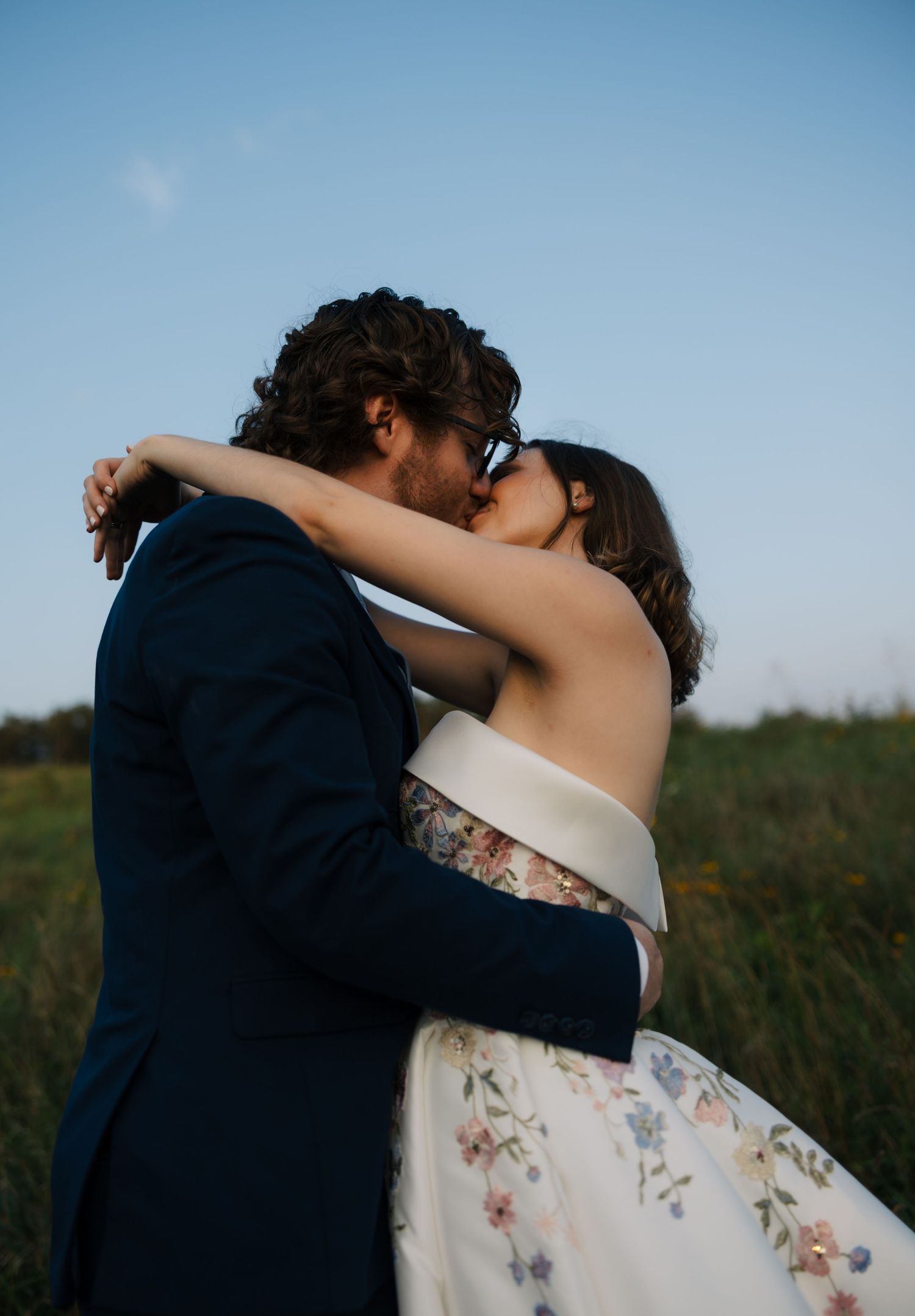 Bride and groom share a passionate kiss outdoors embracing under a clear blue sky.