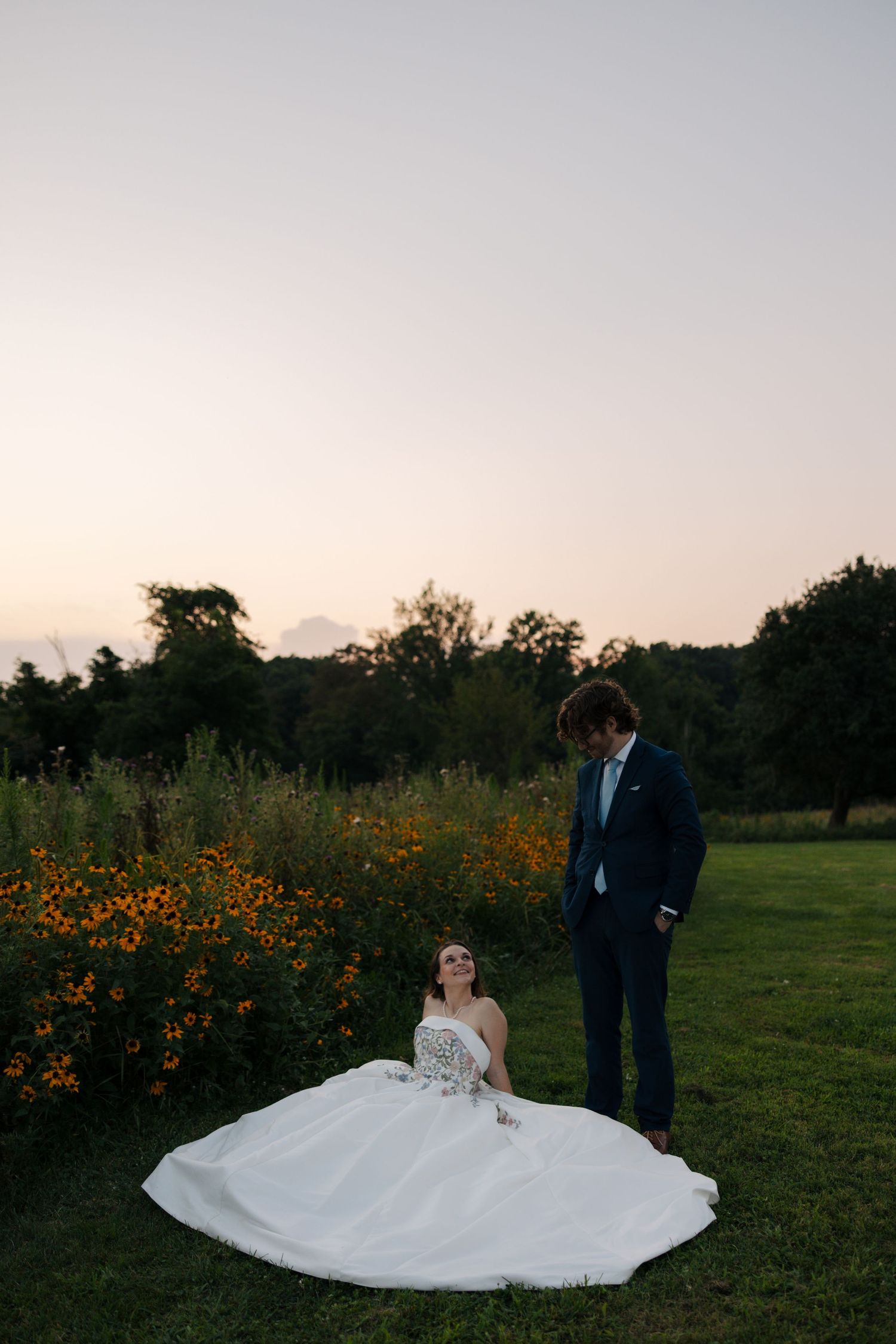Bride in strapless floral ball gown sits in garden as groom in navy suit gazes down at her during sunset.
