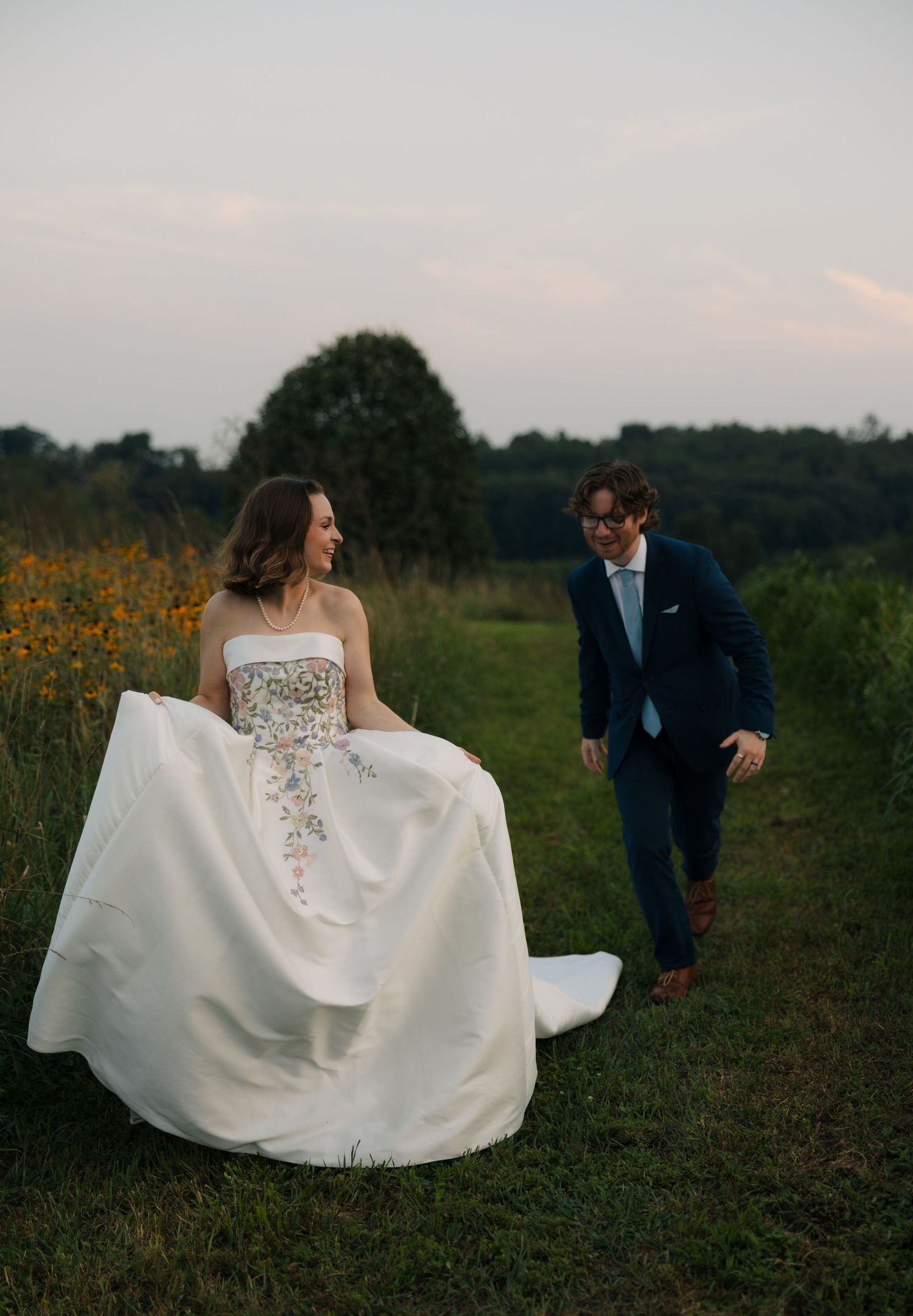 Bride and groom walk through wildflower meadow at sunset, sharing a loving glance in outdoor setting.