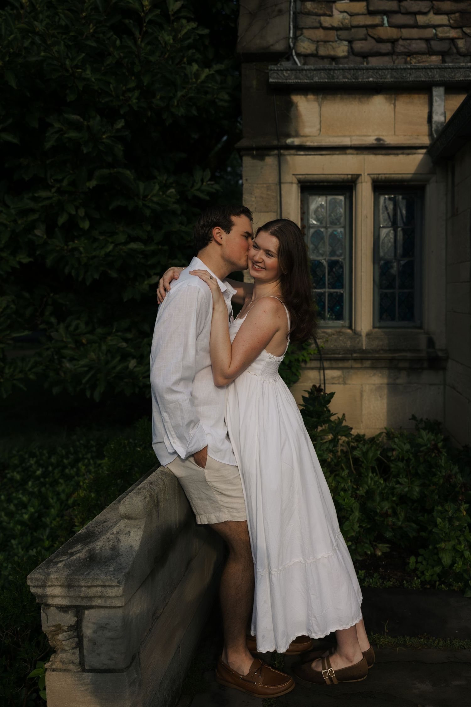 Couple in white outfits sharing a romantic moment by a stone wall outside a classic building surrounded by lush greenery.