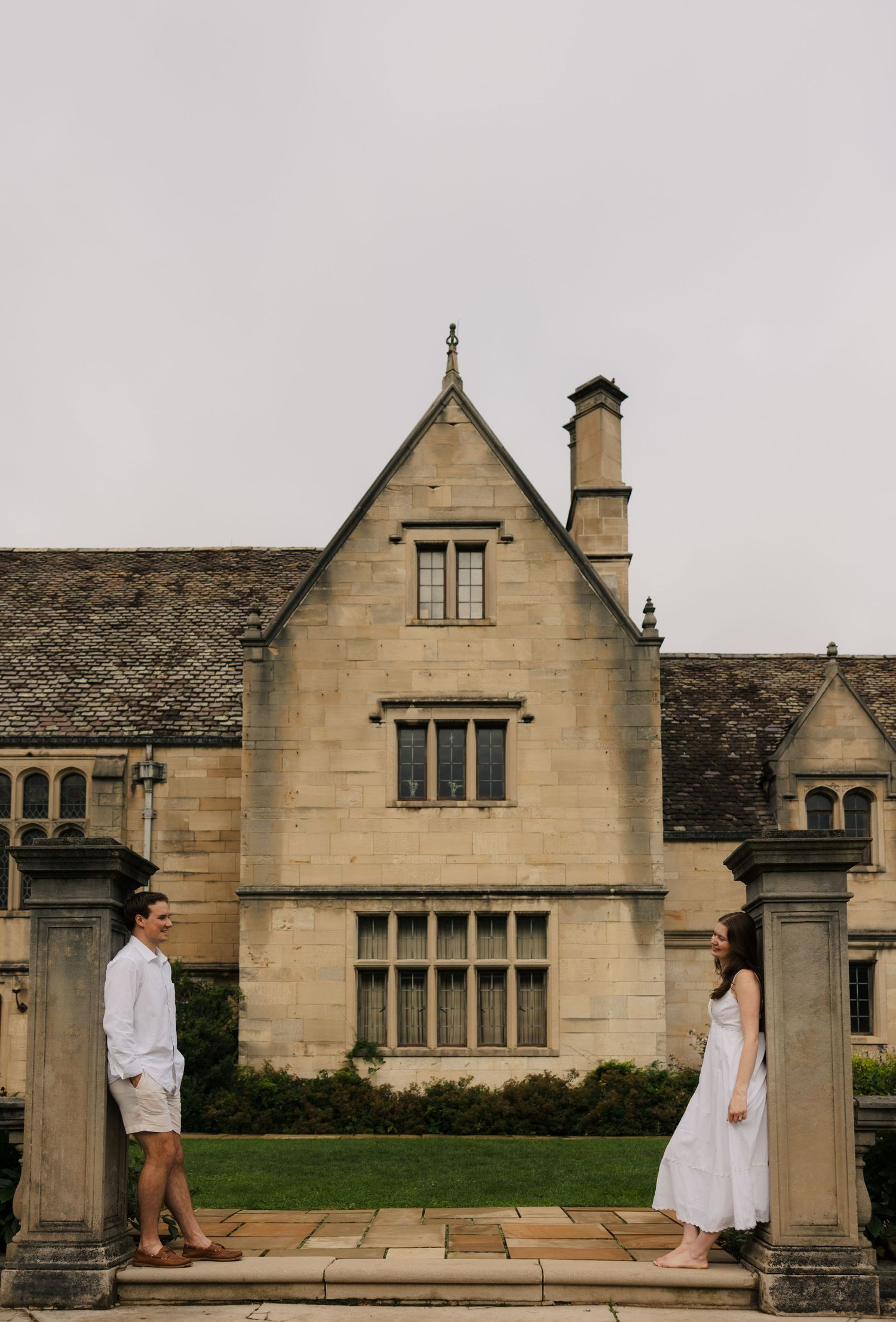 Engaged couple standing apart on stone steps facing each other before a grand historic manor.