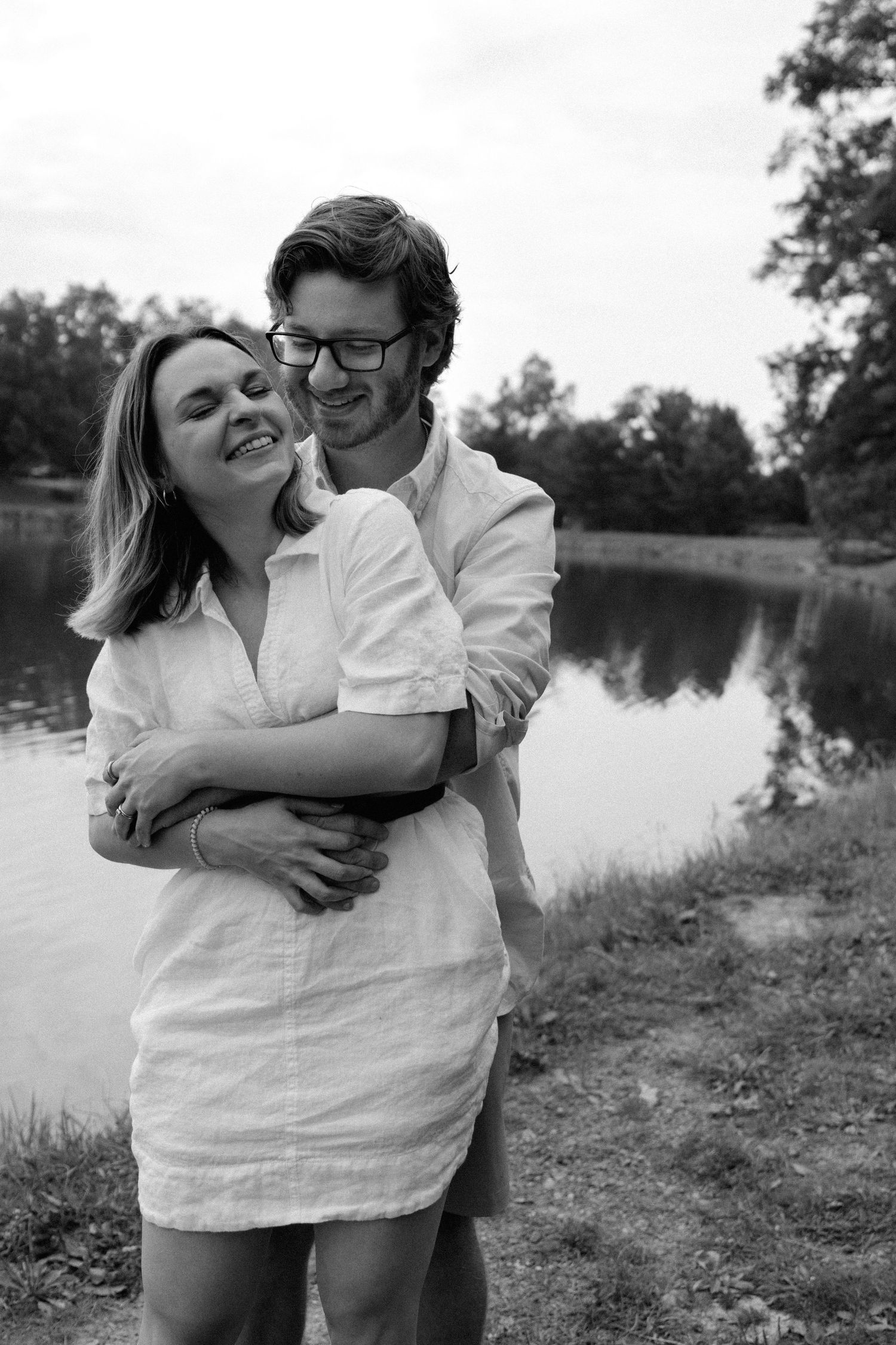 A couple shares a tender embrace while standing near a serene lake in black and white photography.