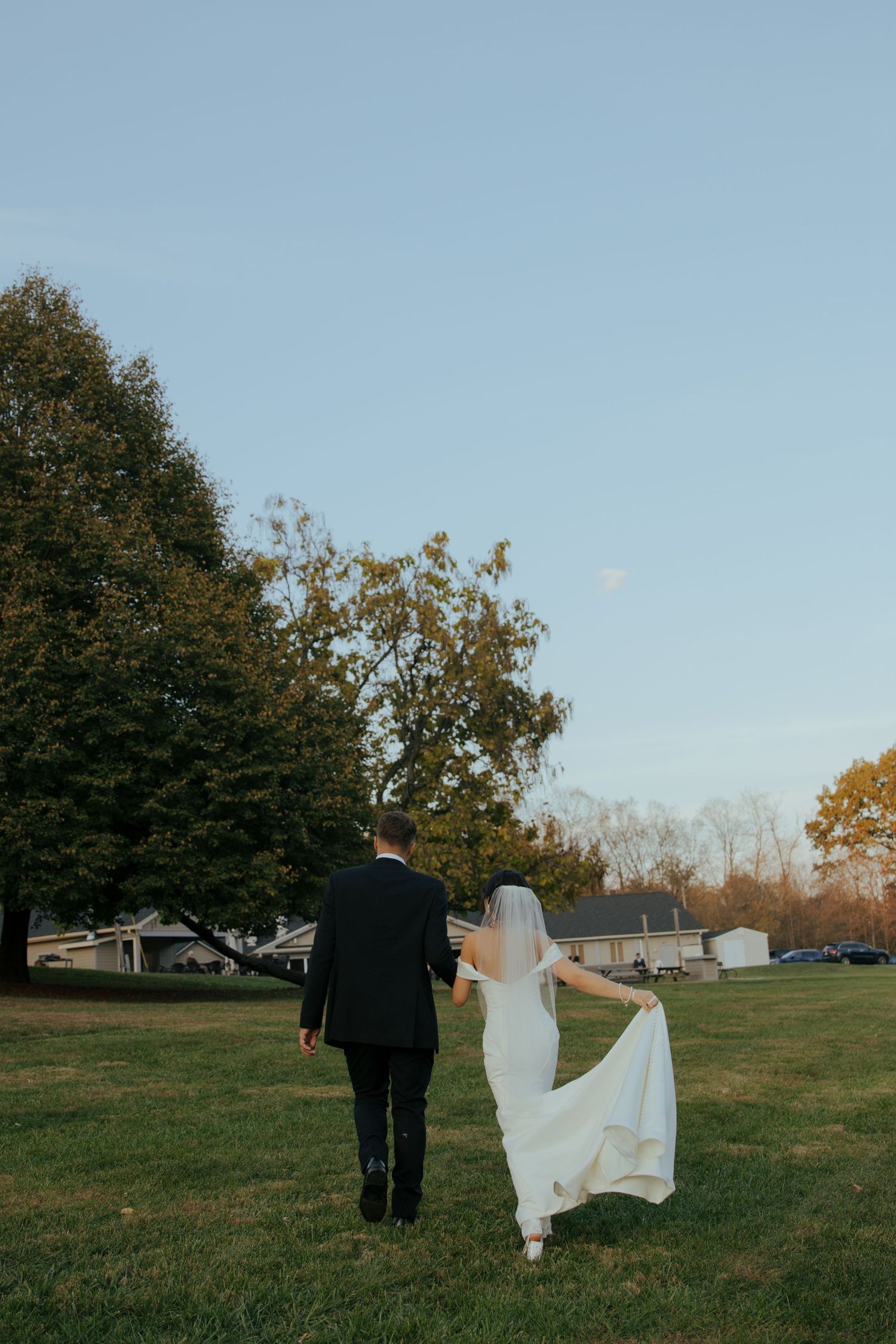 Newlyweds walk together across a grassy field on a clear autumn day.