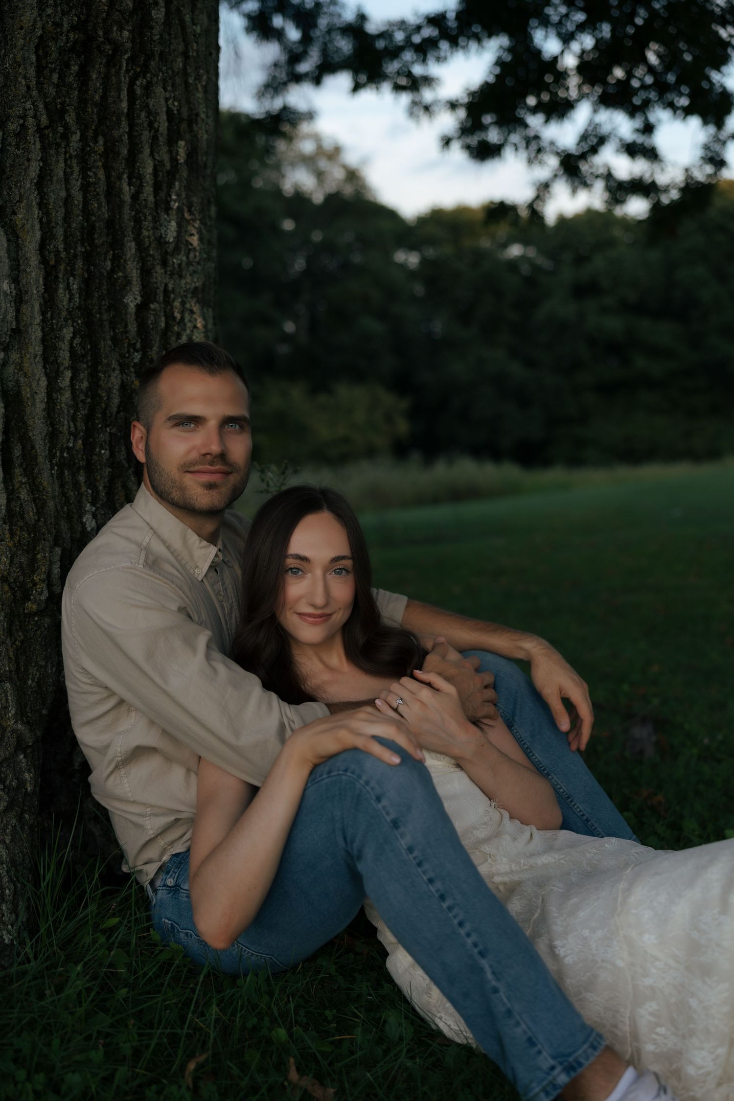 Couple sitting together against tree trunk in park at dusk, bride in white dress.