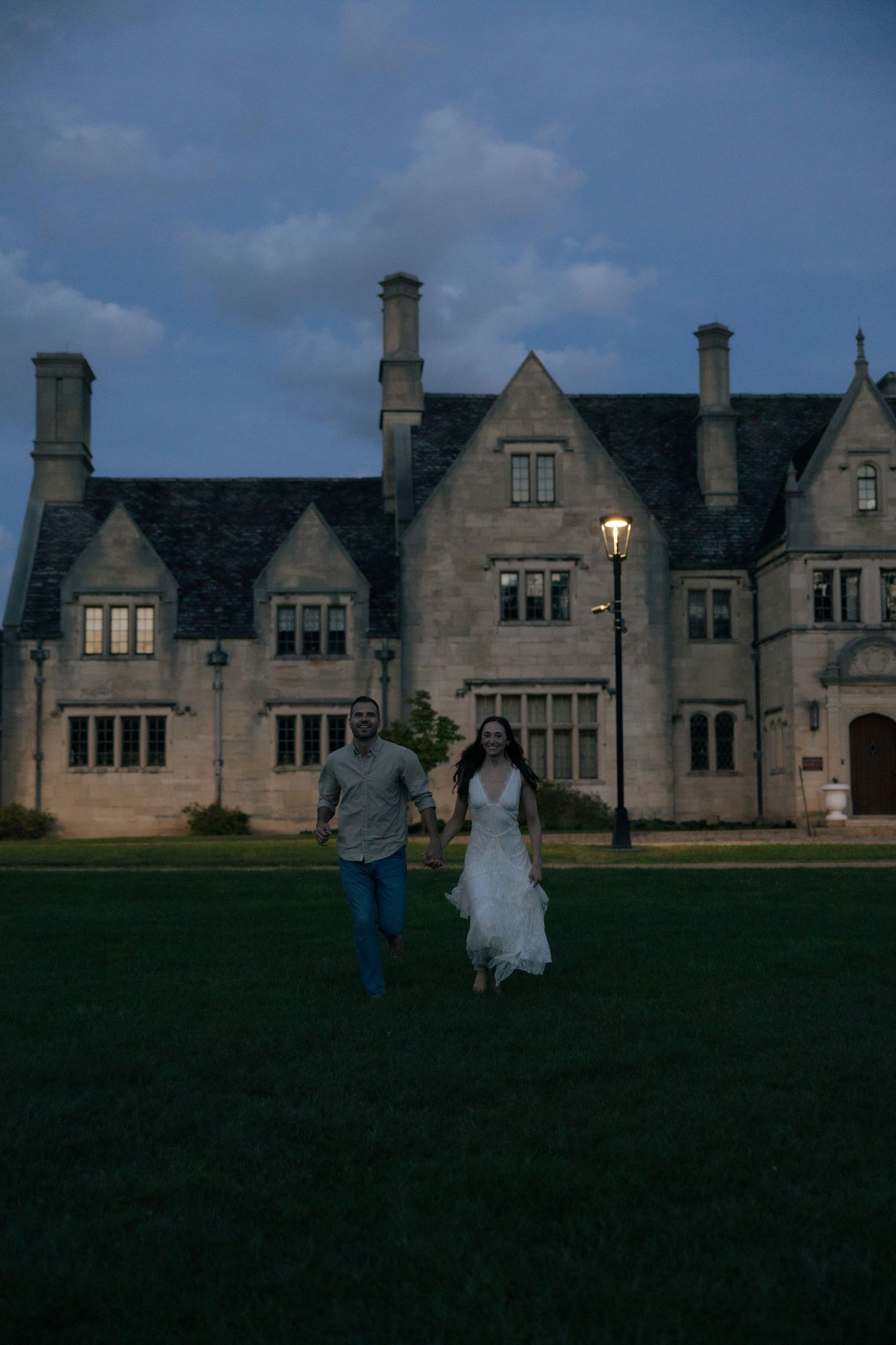 Couple holding hands running across green lawn in front of elegant stone manor at dusk.