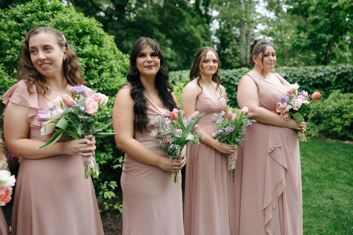 Four bridesmaids in blush pink gowns holding colorful bouquets stand outdoors surrounded by lush green foliage.