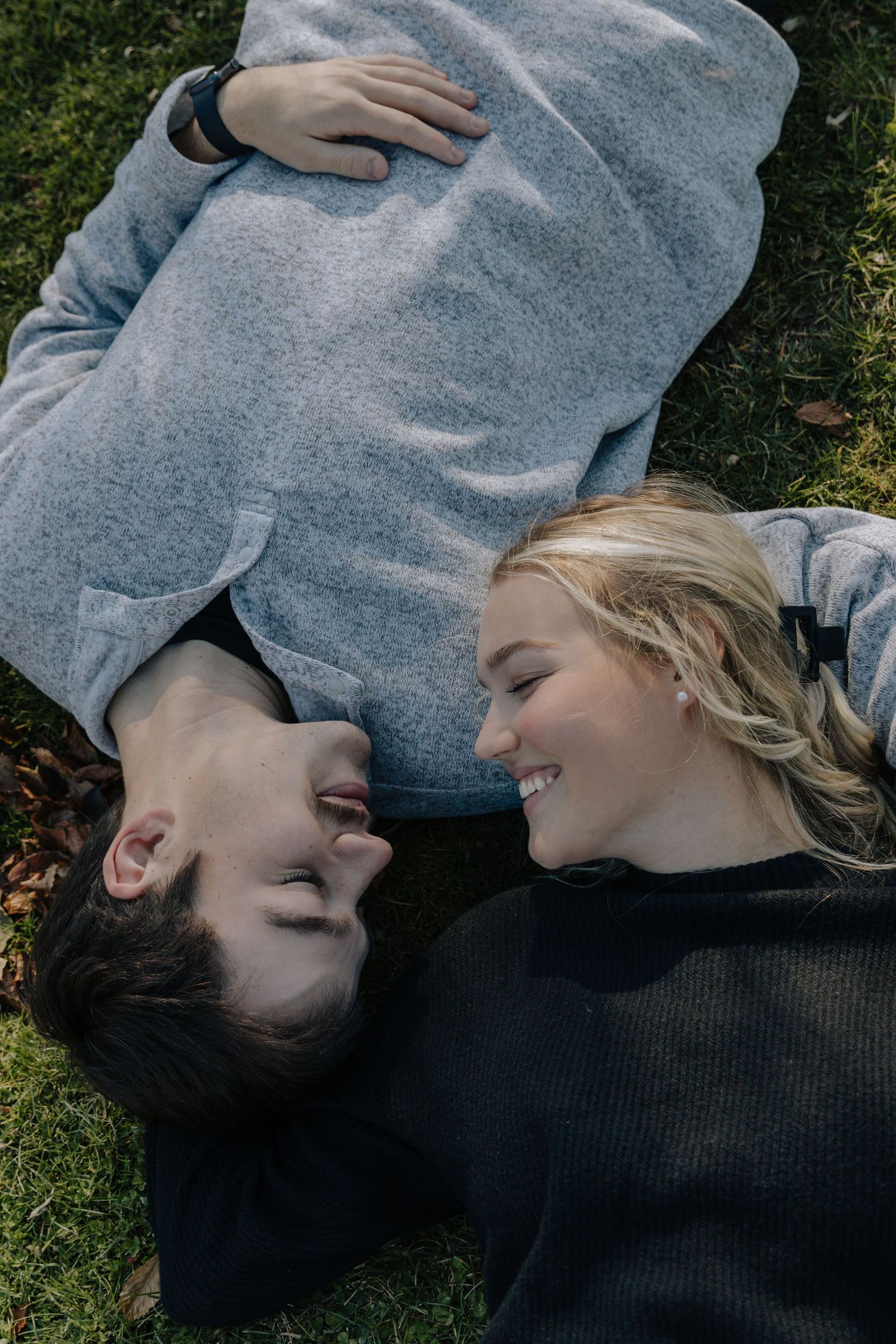 Two people lying on grass wearing cozy sweaters while sharing a tender moment together.