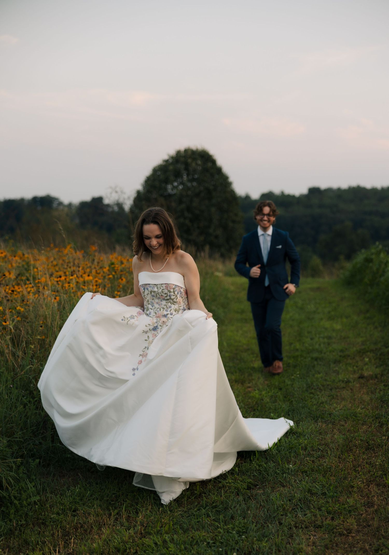 Joyful bride lifts white floral wedding dress while groom in navy suit runs toward her through meadow.