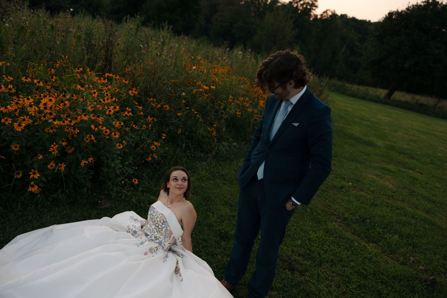 Bride in white floral gown sits on grass looking up at groom in navy suit near orange flowers.
