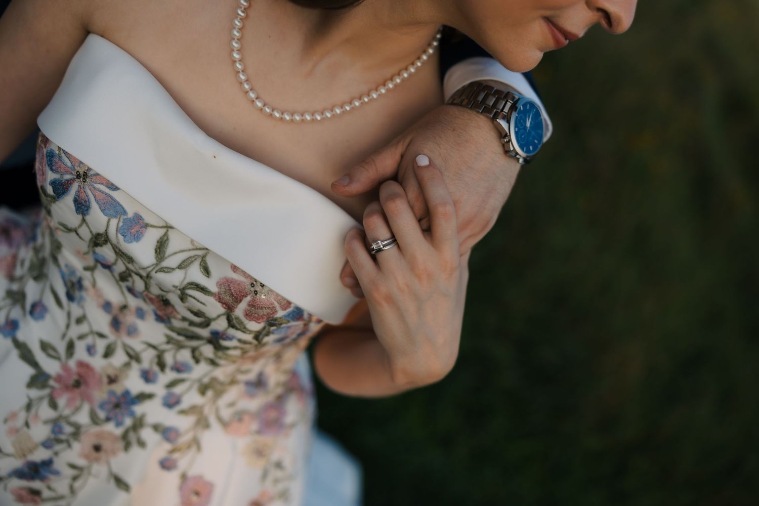 Close-up of bride in floral strapless gown with pearl necklace, holding groom's arm with engagement ring visible.