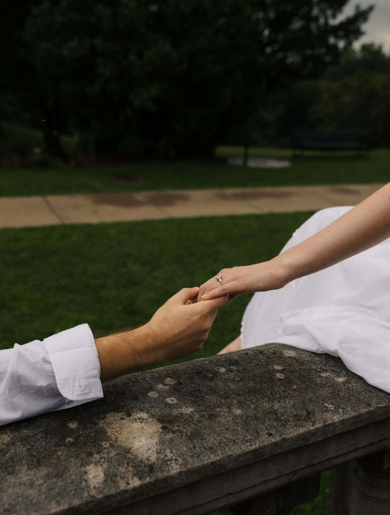 Close-up of engaged couple holding hands on stone ledge, woman wearing engagement ring in white dress.