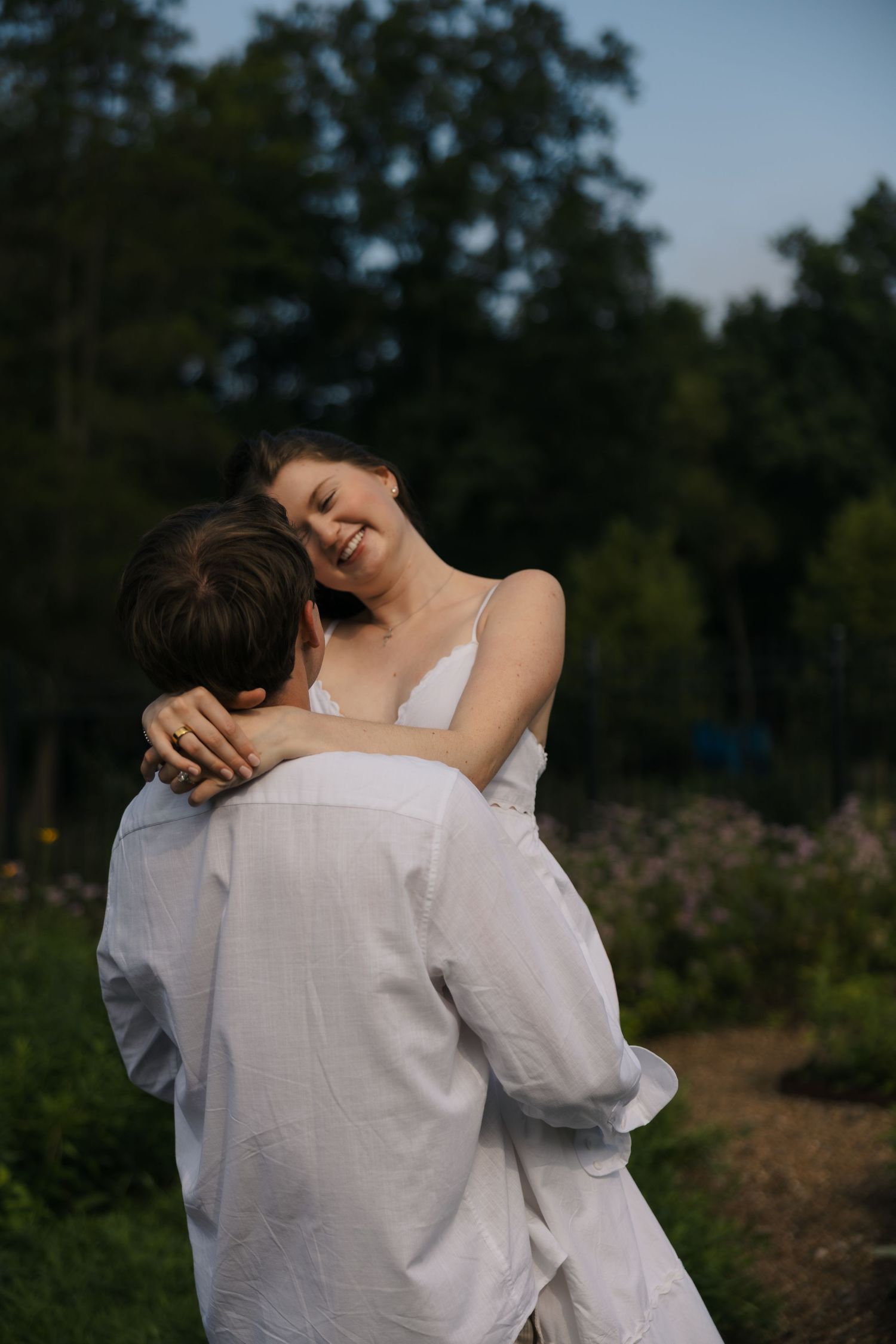 Man lifting smiling woman in white dress outdoors at golden hour.