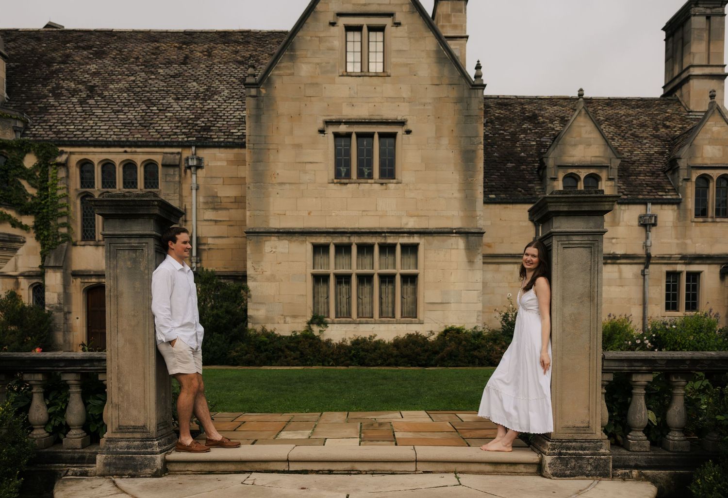 Engaged couple standing apart between stone pillars before a grand historic English manor house.