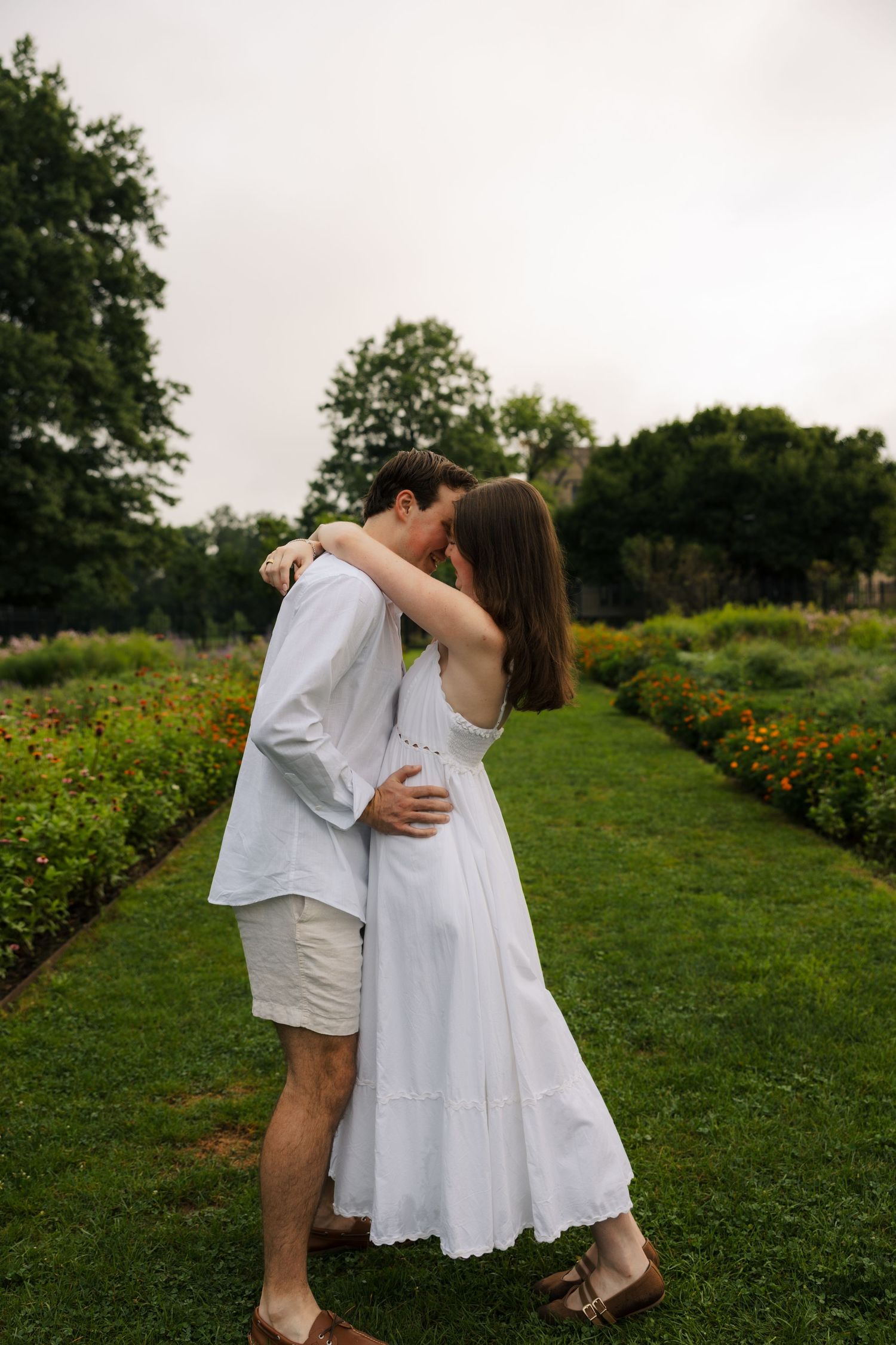 A couple embraces and kisses in a lush garden path, wearing white outfits, surrounded by colorful flowers and green trees.