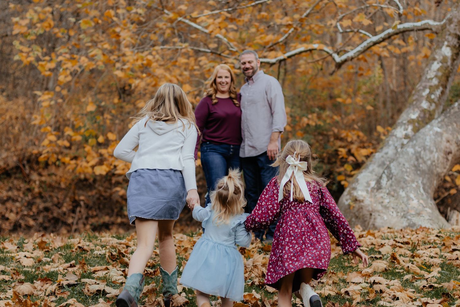Family of five walking through autumn forest with golden leaves, parents and three daughters enjoying fall outdoor photo session.